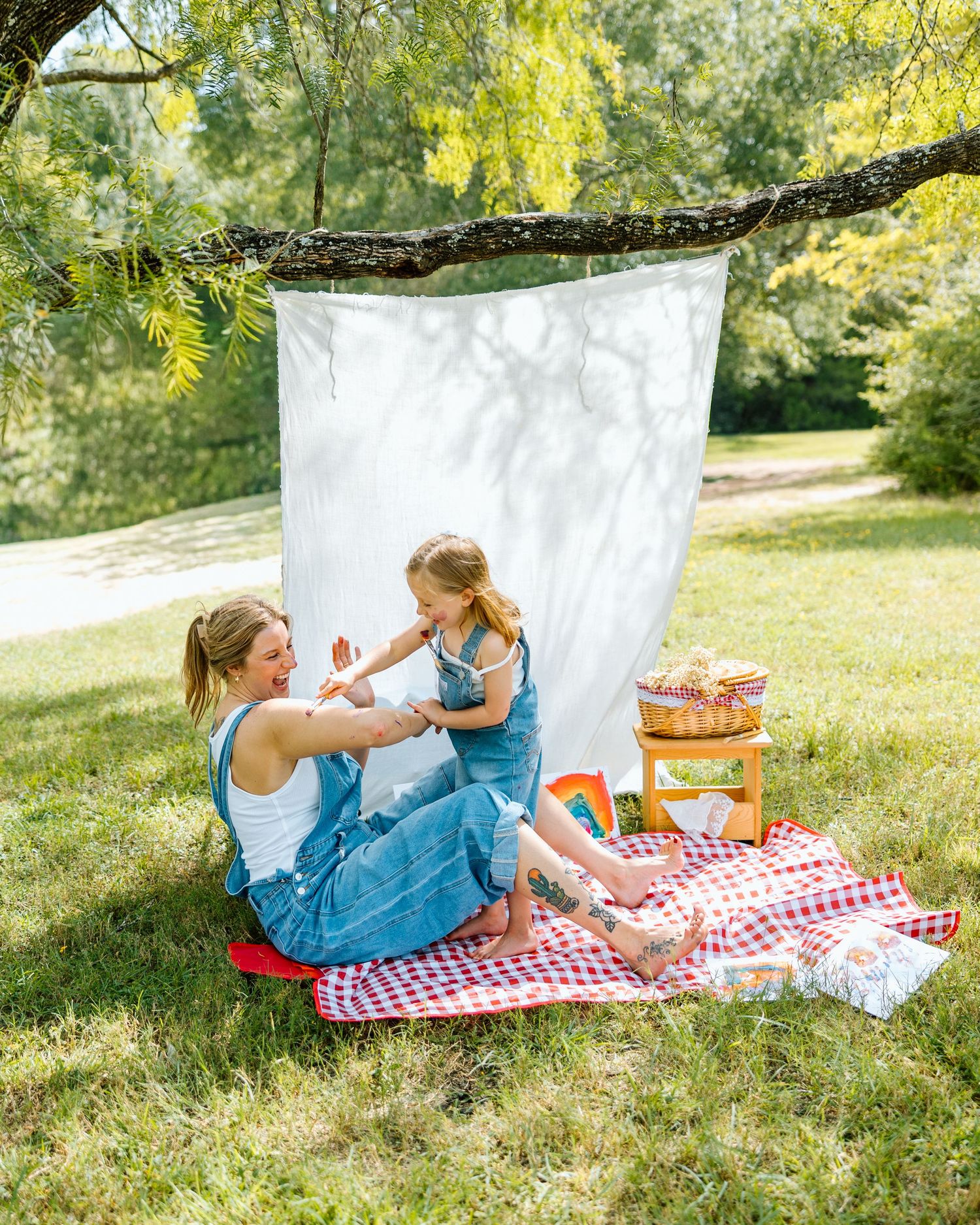 Mom and daughter in denim overalls enjoy a sunny outdoor picnic on a red checkered blanket with a white backdrop.