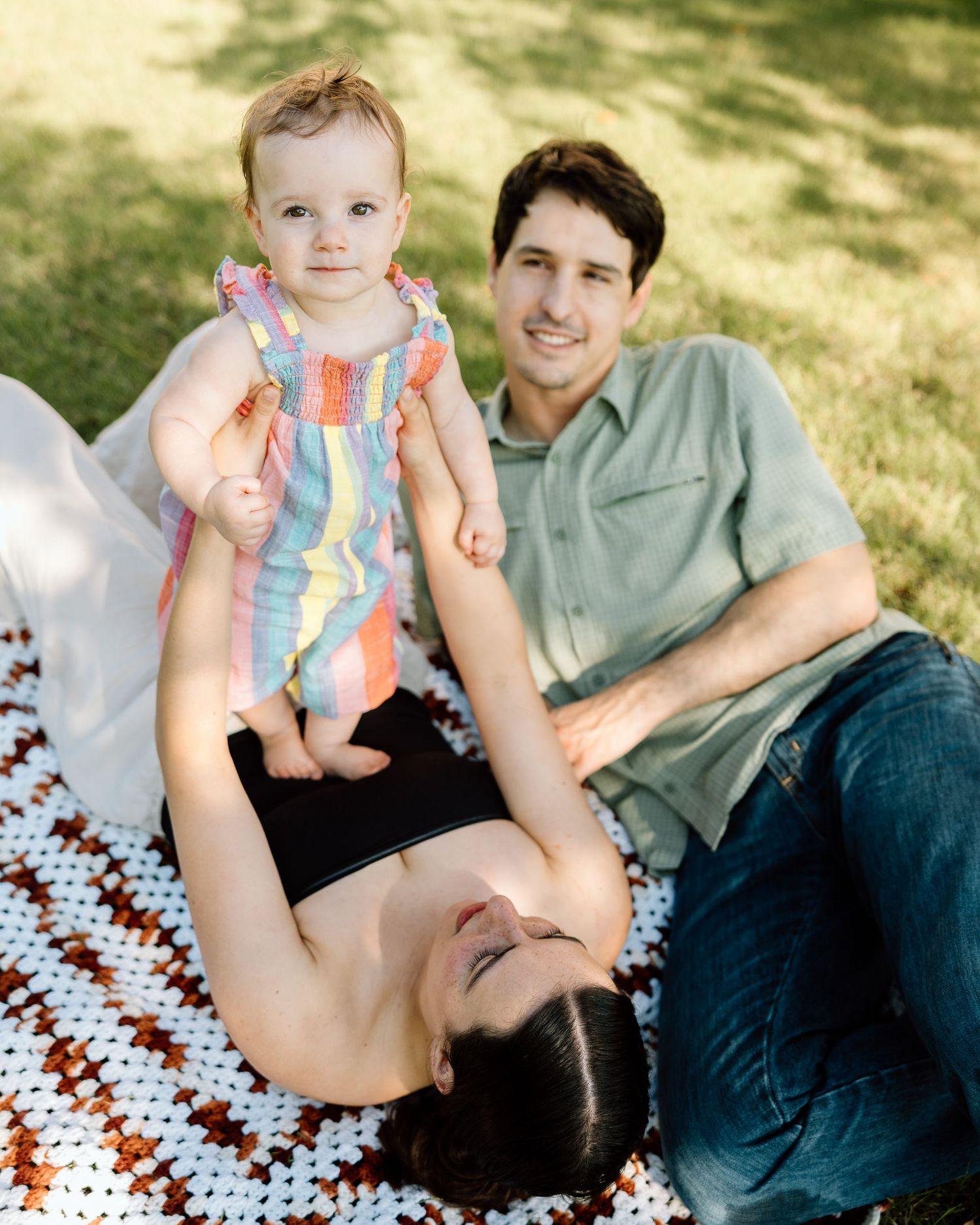 A young family relaxes on a crochet blanket outdoors, with parents smiling at their baby girl in a colorful striped romper.