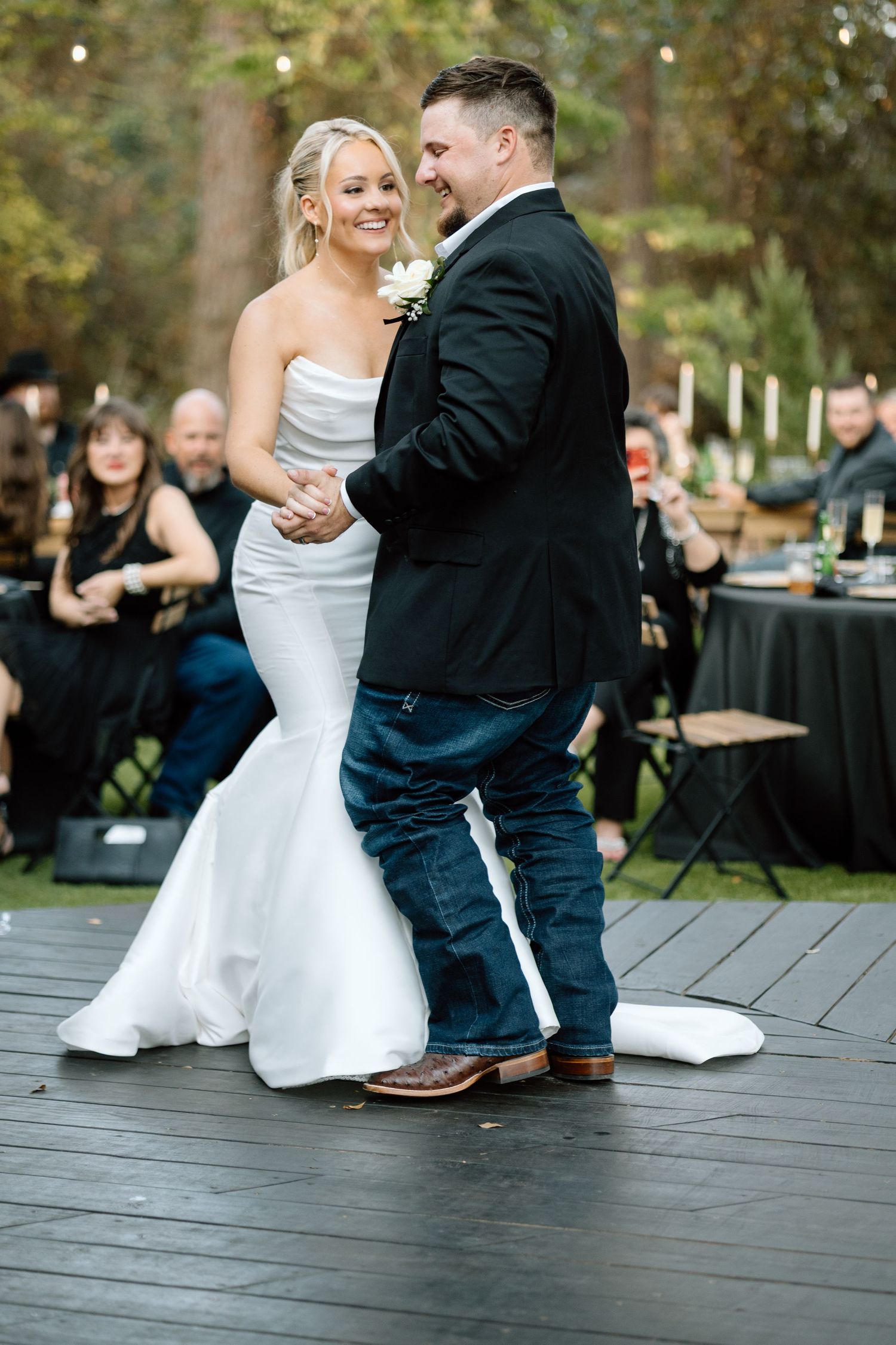 Smiling bride and groom share first dance on outdoor wooden dance floor surrounded by wedding guests.