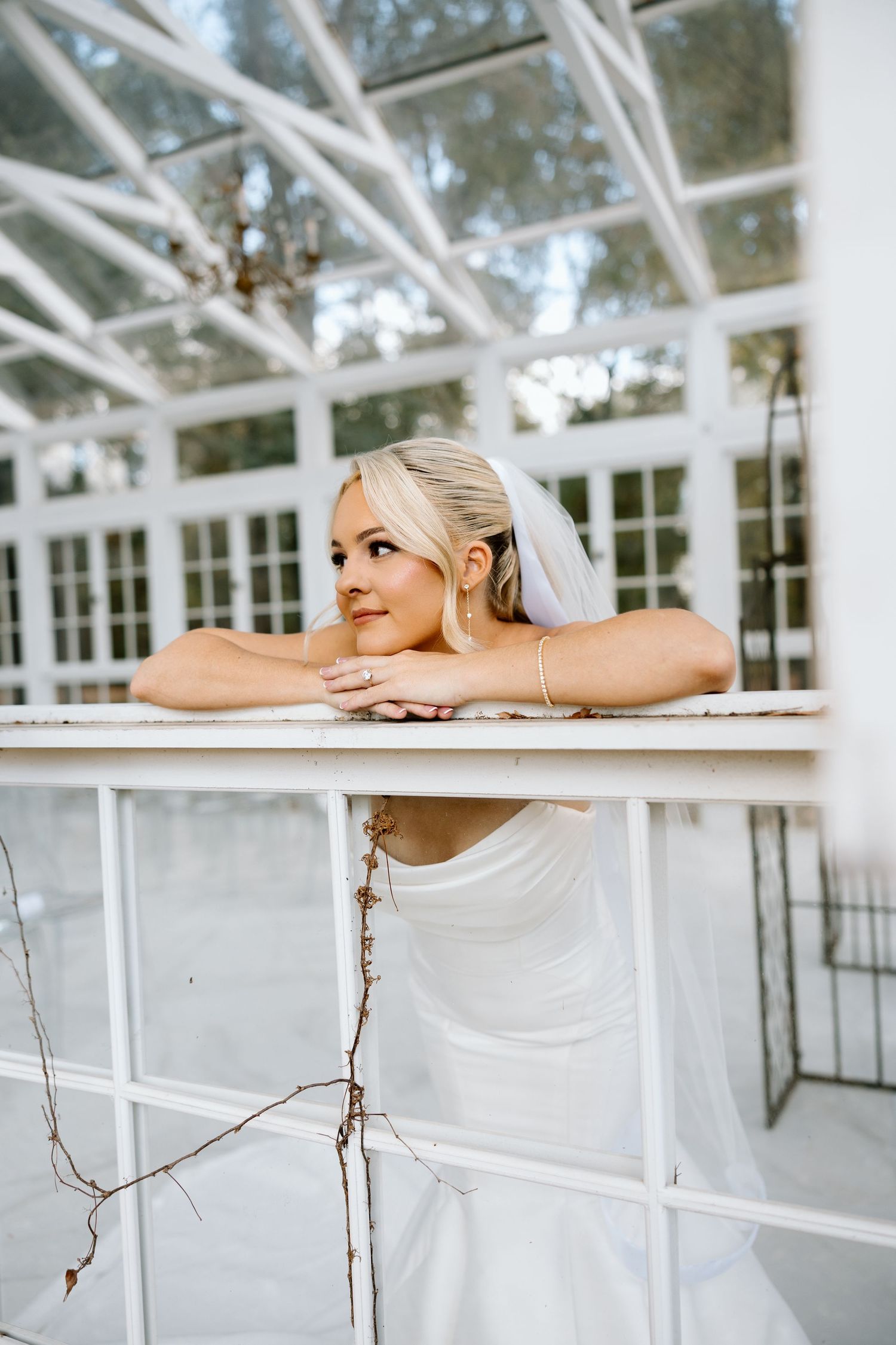 Elegant bride in strapless white gown and veil leans on white railing inside a sunlit greenhouse conservatory.