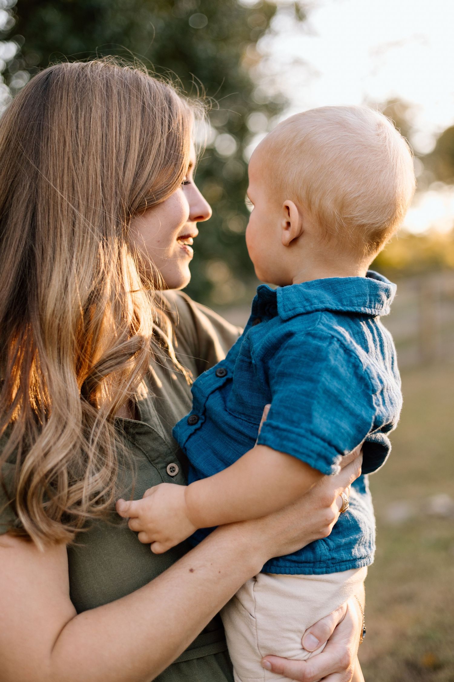 A mother holds her baby boy close while smiling at him outdoors in golden hour light.