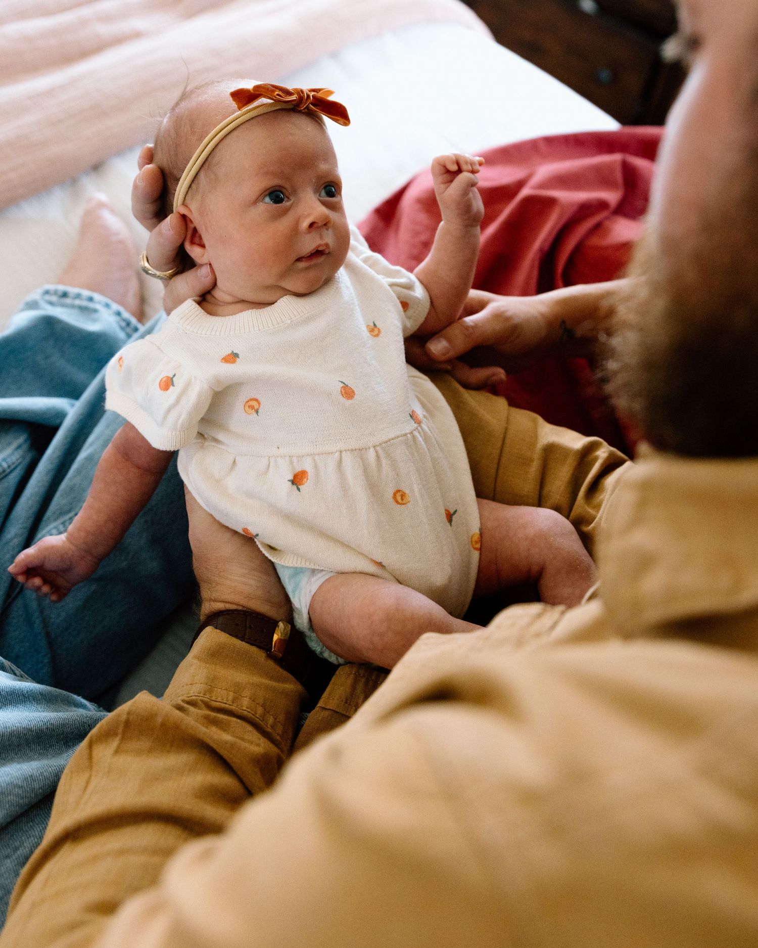 Adorable newborn baby girl in white floral onesie and orange bow headband looks up at her father.