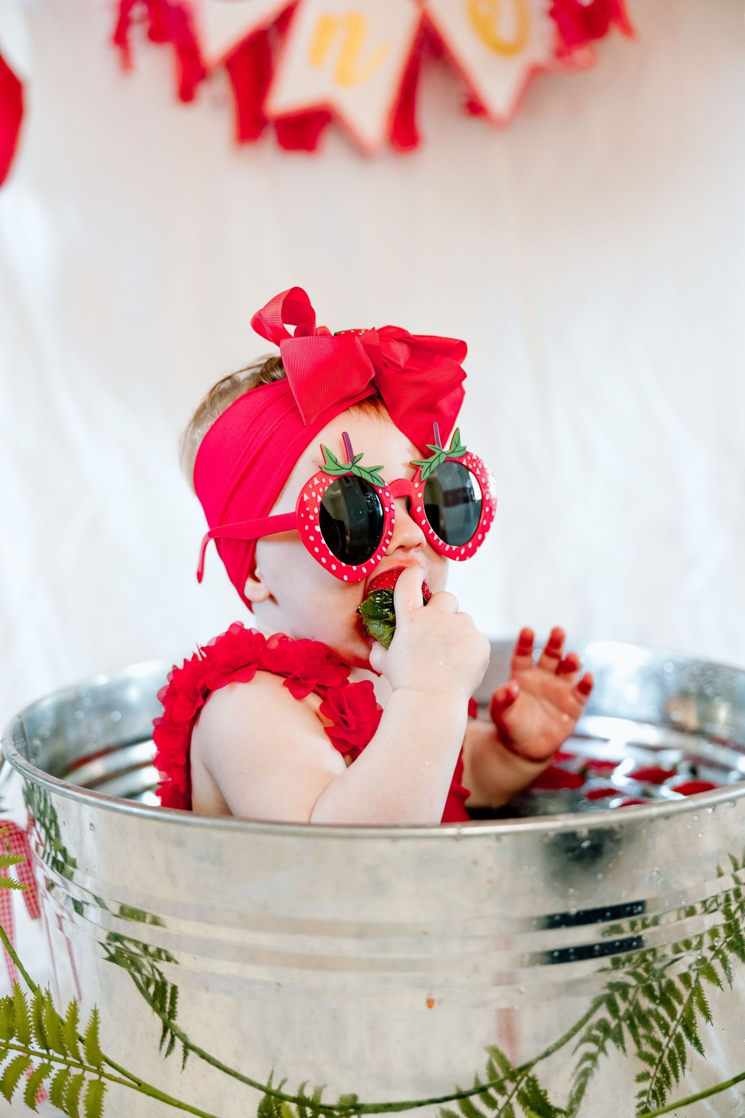 Baby girl in red ruffle swimsuit and strawberry sunglasses sitting in galvanized tub at first birthday party.