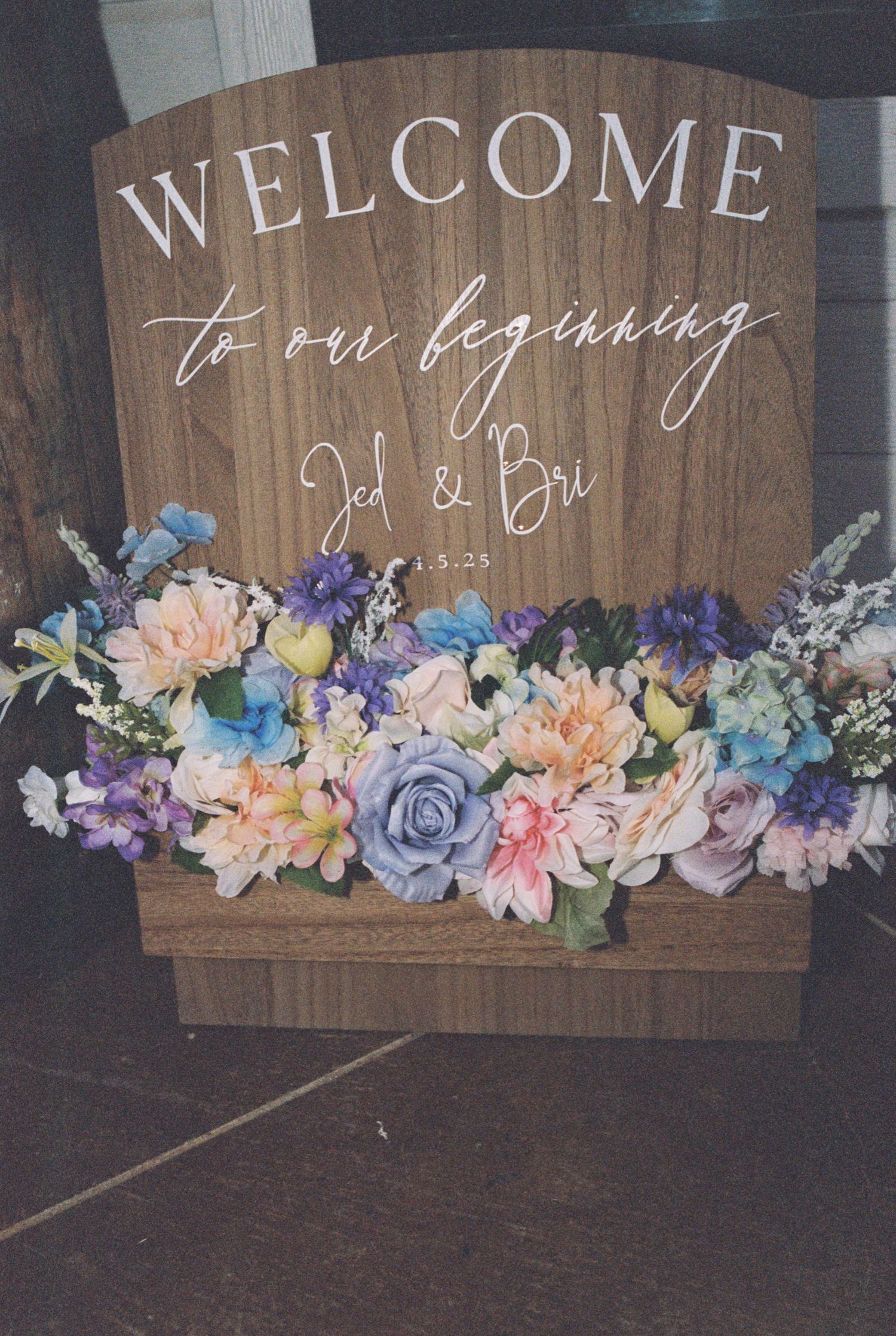 A rustic wooden welcome sign decorated with pink, blue and purple flowers for a wedding.