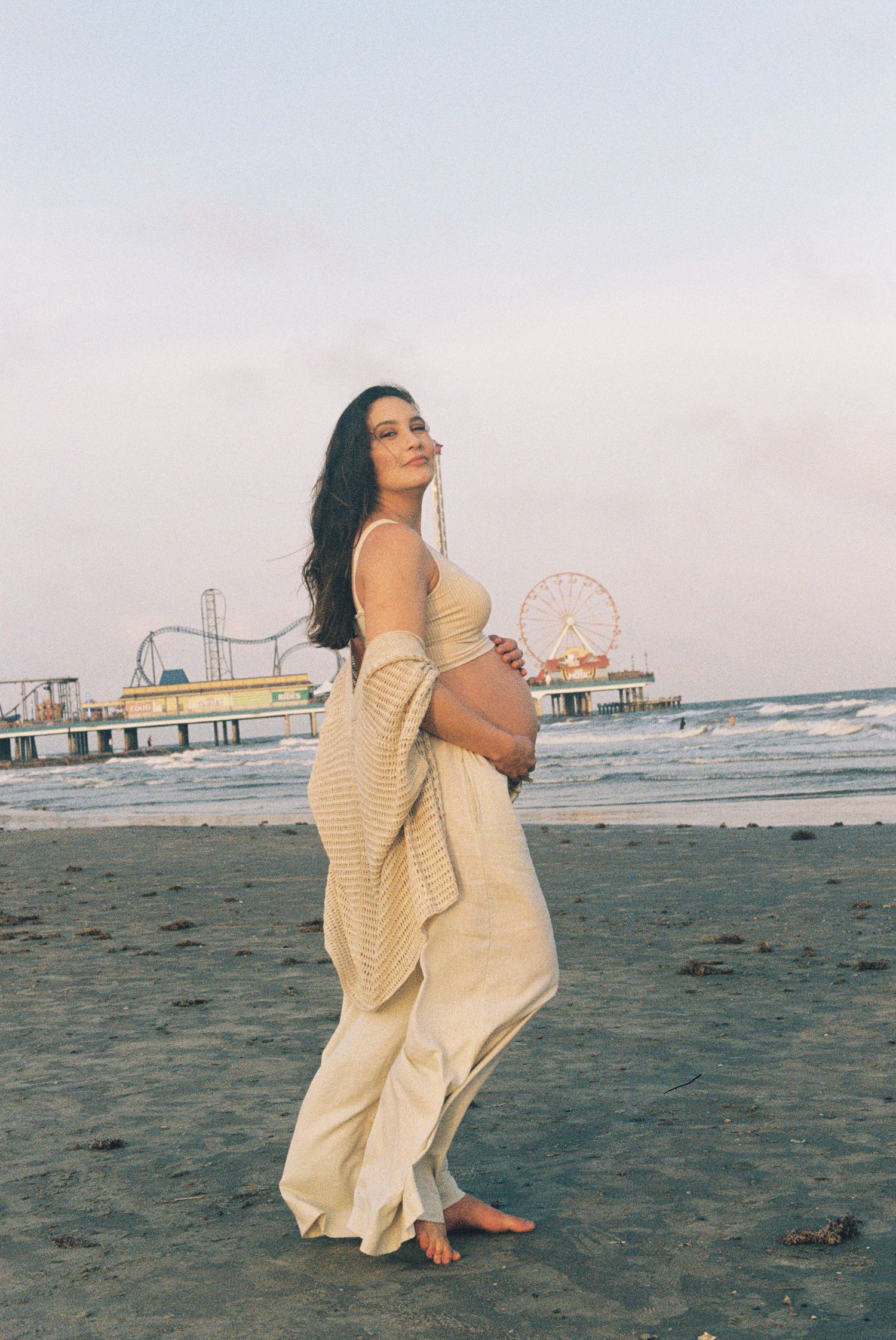 A pregnant woman in a flowing cream dress poses on the beach with an amusement park backdrop.