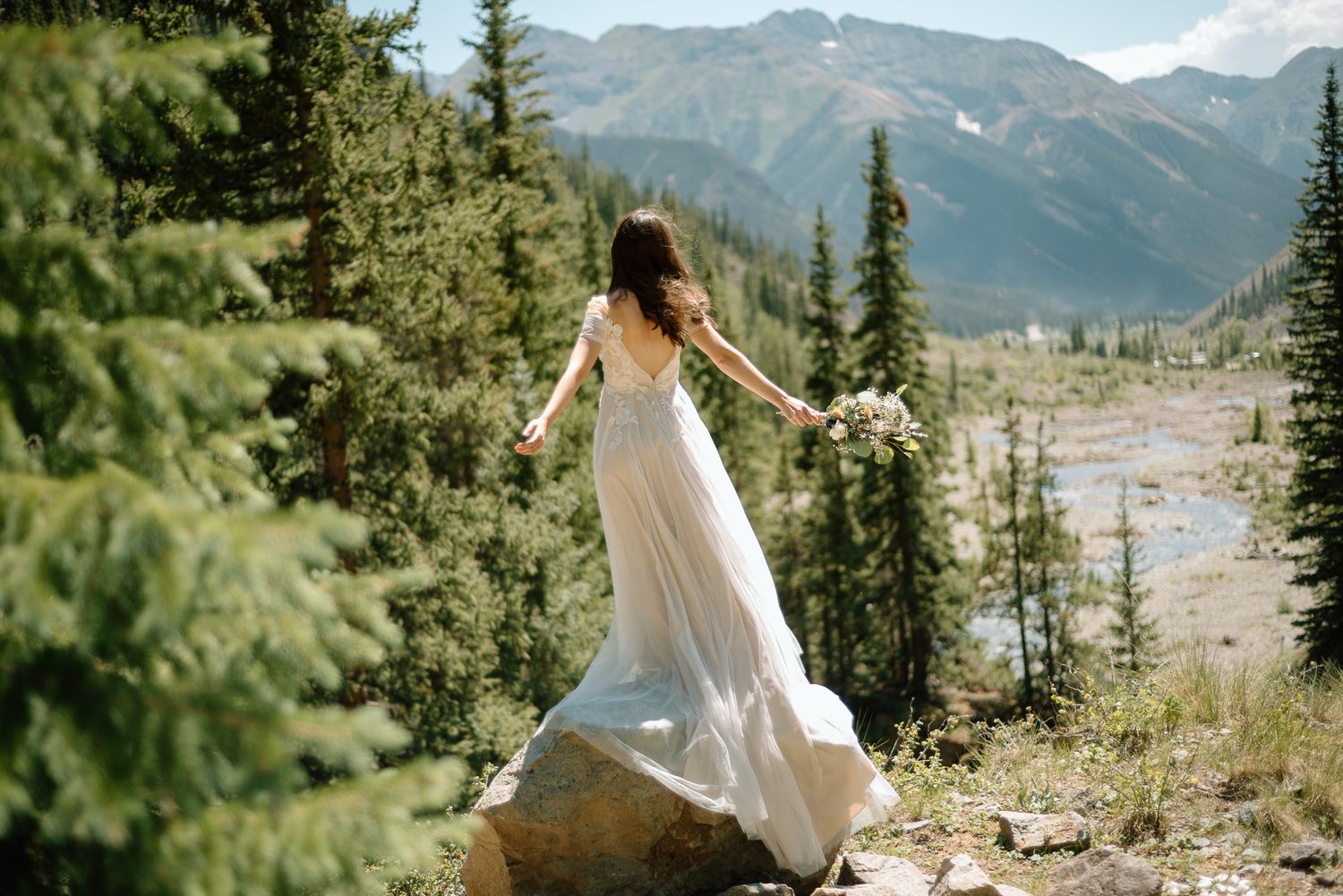 Bride in flowing gown twirls on mountain peak with scenic valley view.