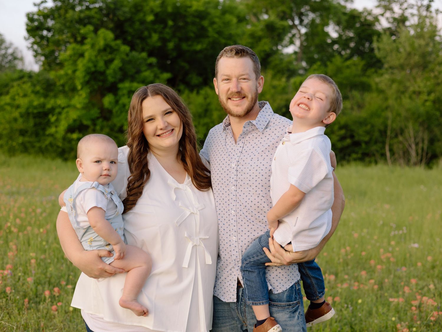 A family of four stands together in a sunlit meadow with green trees in the background.