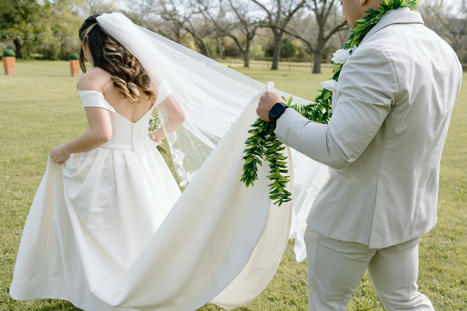 A couple in wedding attire stands on a grassy lawn as the groom helps adjust the bride's flowing white dress and veil.