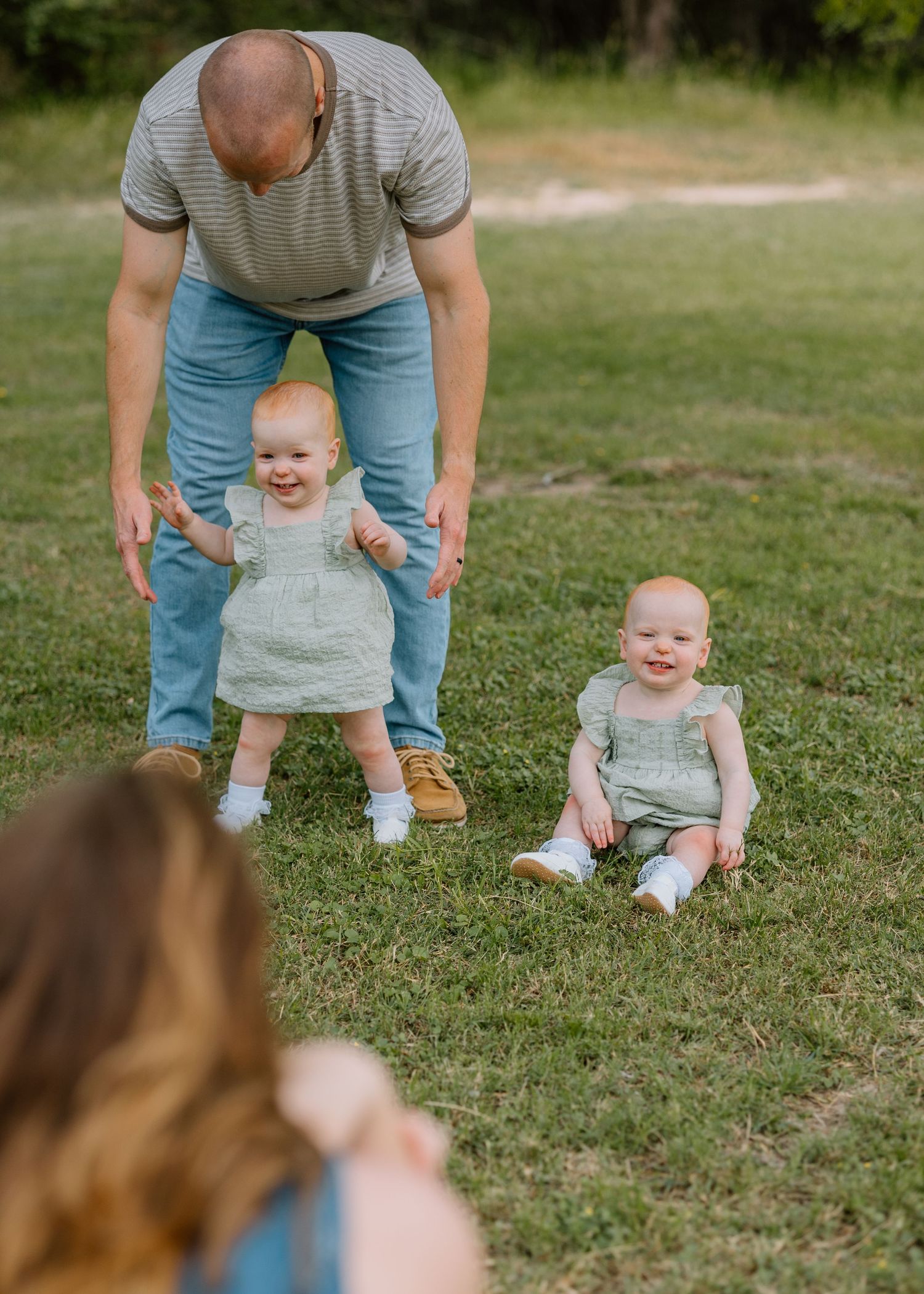 Adult helps toddlers practice walking on green grass during outdoor playtime in casual summer setting.
