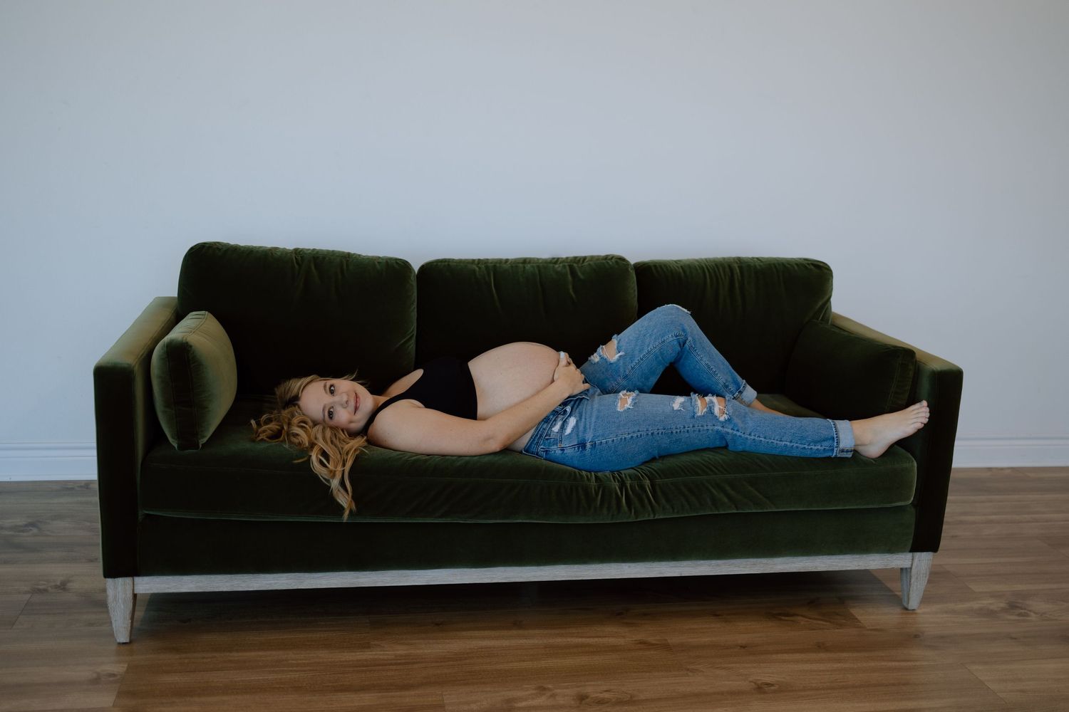 A person in ripped jeans and a black top lies asleep on a dark green velvet couch against a white wall.