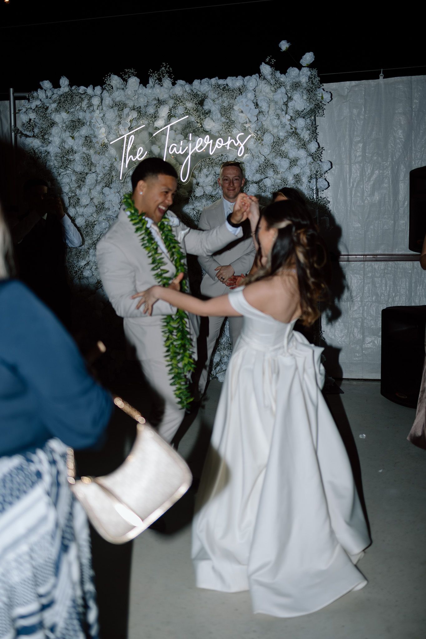 Couple dances at reception in front of white floral wall with neon signage.