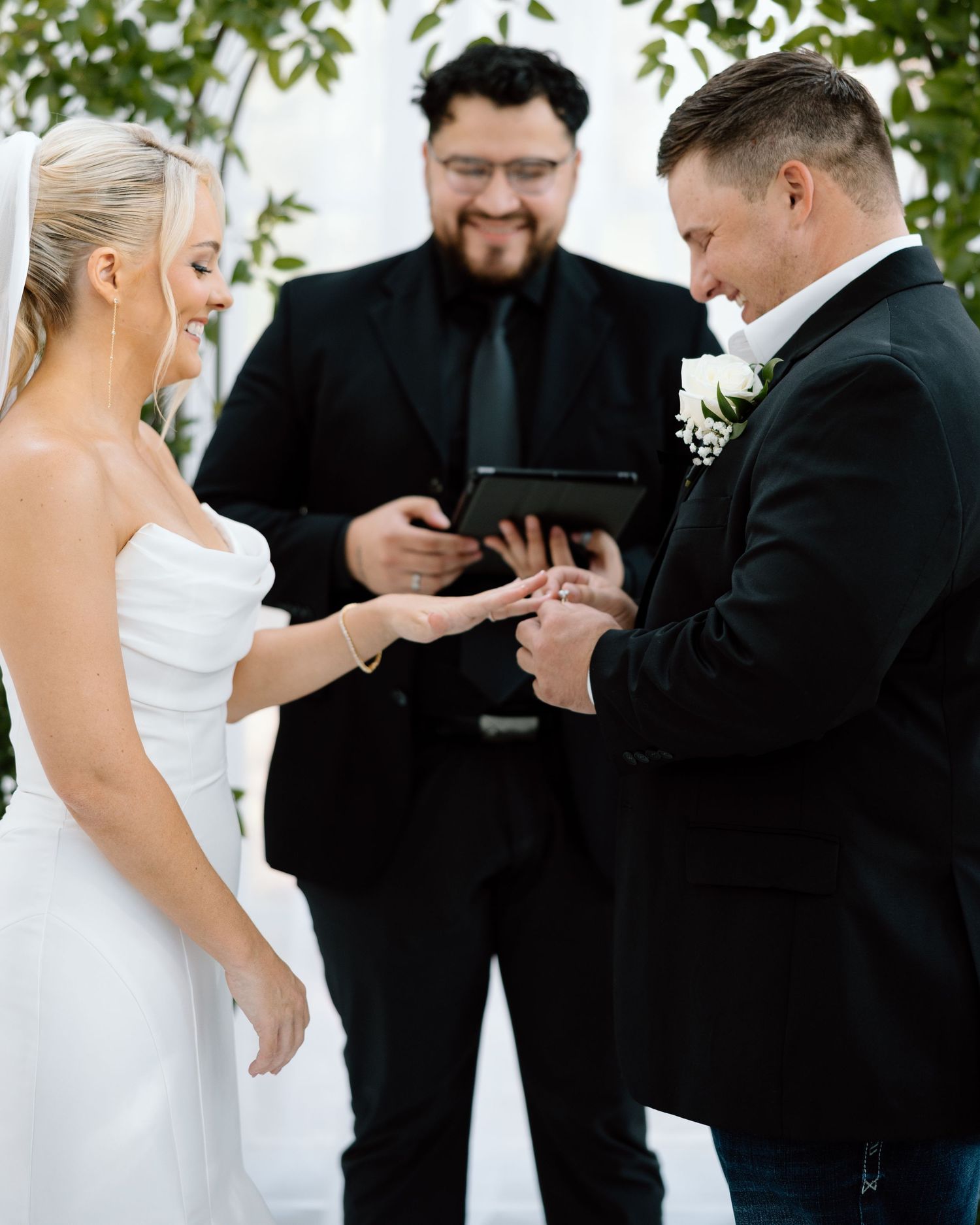 Groom places wedding ring on brides finger during outdoor ceremony as officiant reads from tablet behind them.
