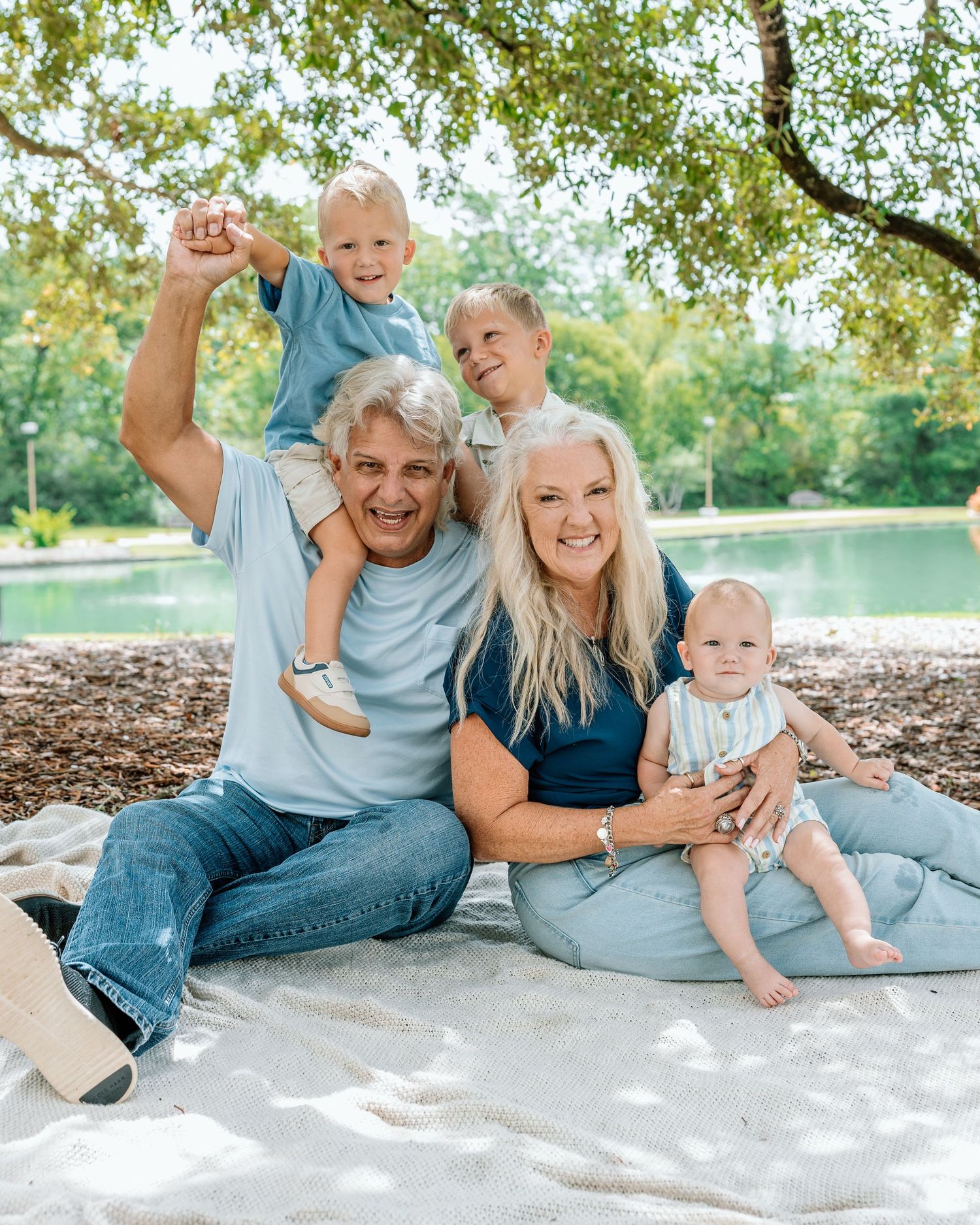 Happy family of five posing outdoors near a pond, with grandparents holding three young children on a white blanket.
