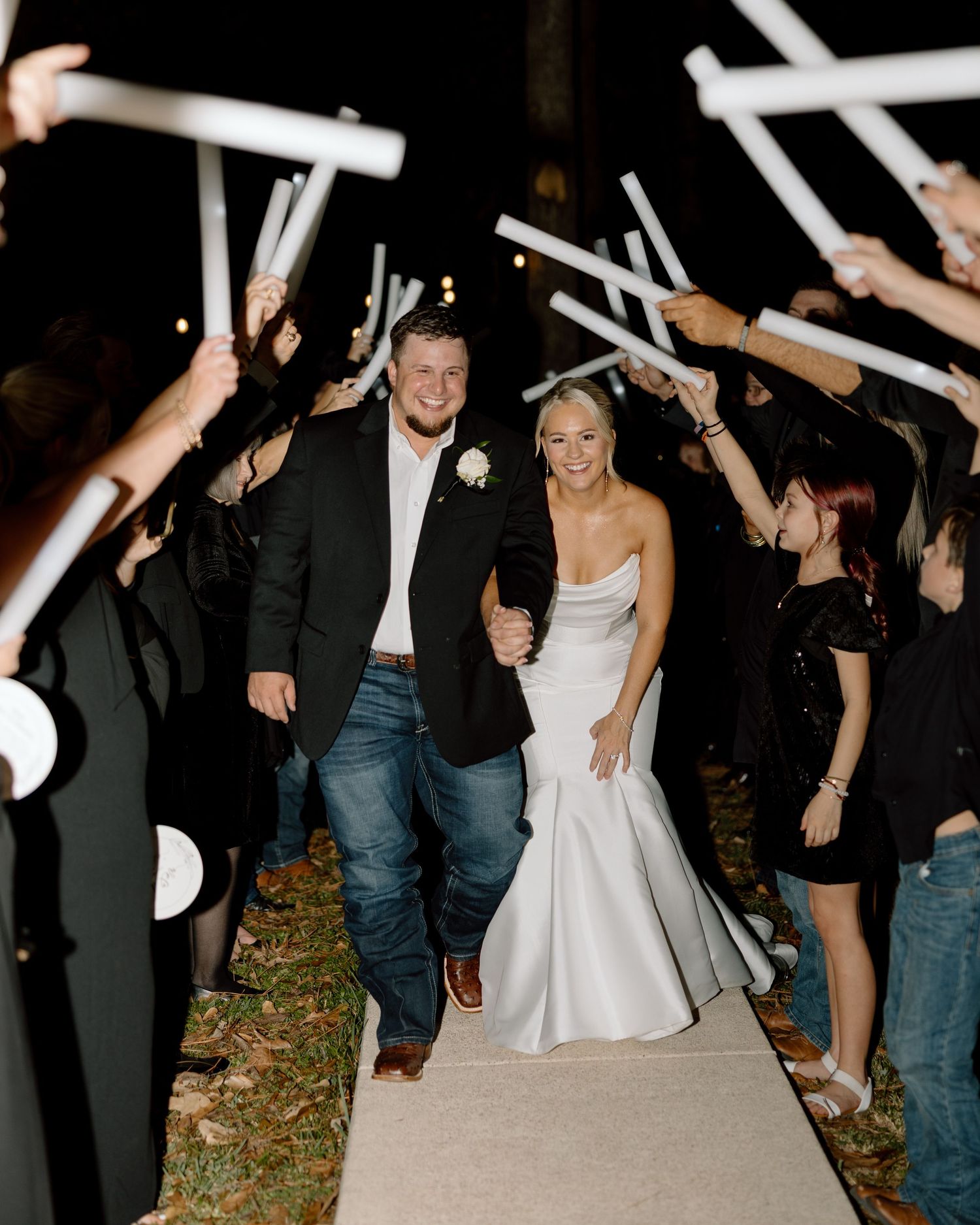 Bride and groom exit wedding reception through cheering guests holding glowing white light wands at night.