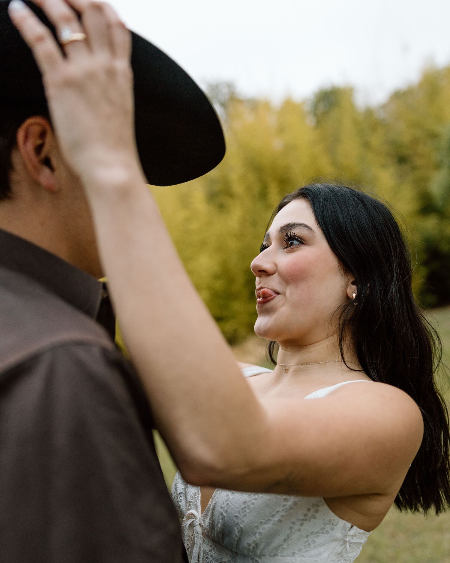 Woman in white dress placing black cowboy hat on mans head outdoors during fall engagement photo session.