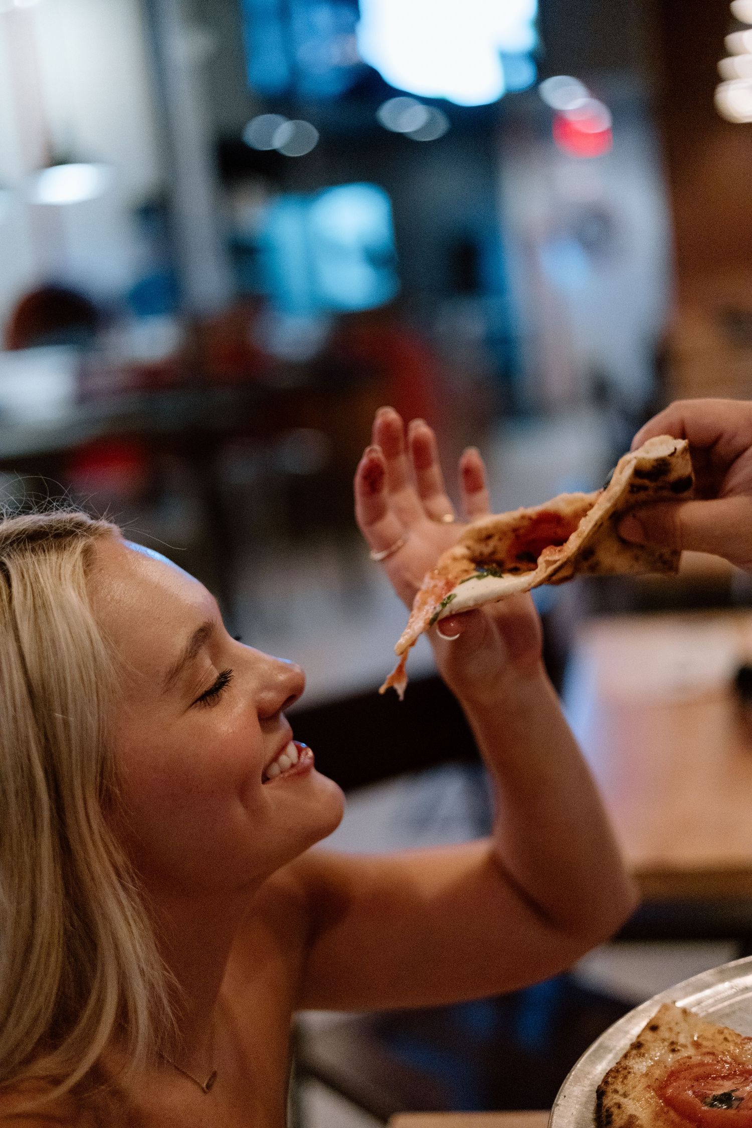 Smiling blonde woman reaching for a slice of pizza at a restaurant, enjoying a casual dining experience.