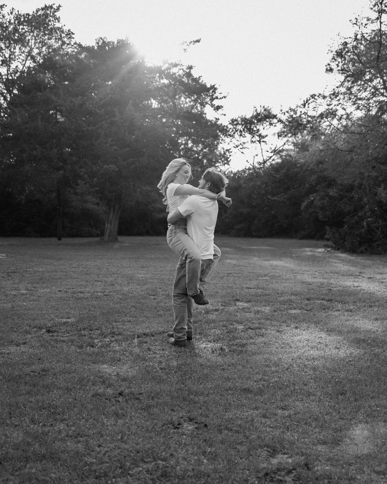 Black and white photo of a couple embracing in a sunlit park, man lifting woman joyfully among lush trees.