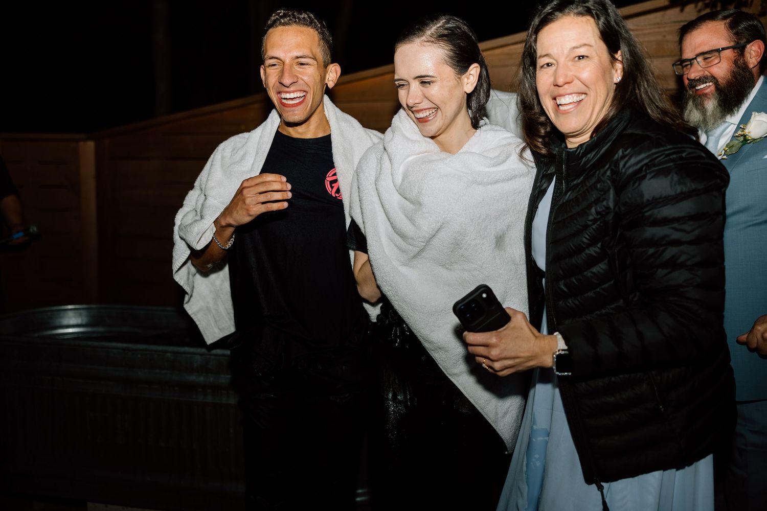 Bride laughing with groom and guest wrapped in blankets at outdoor evening reception.