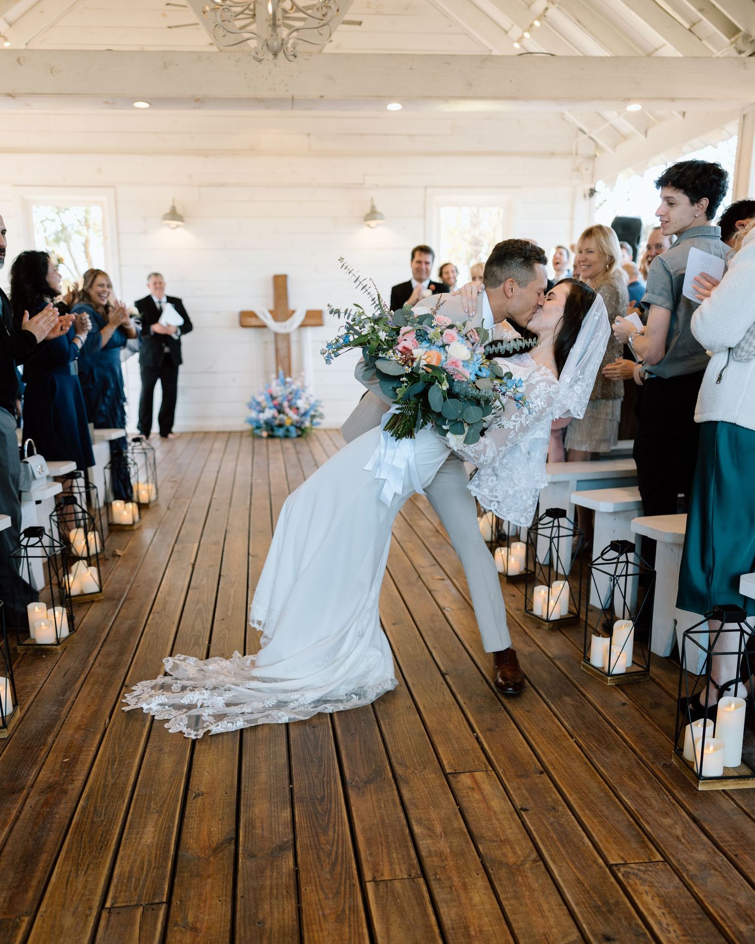 Bride and groom share romantic dip kiss in rustic chapel aisle surrounded by candlelit lanterns.