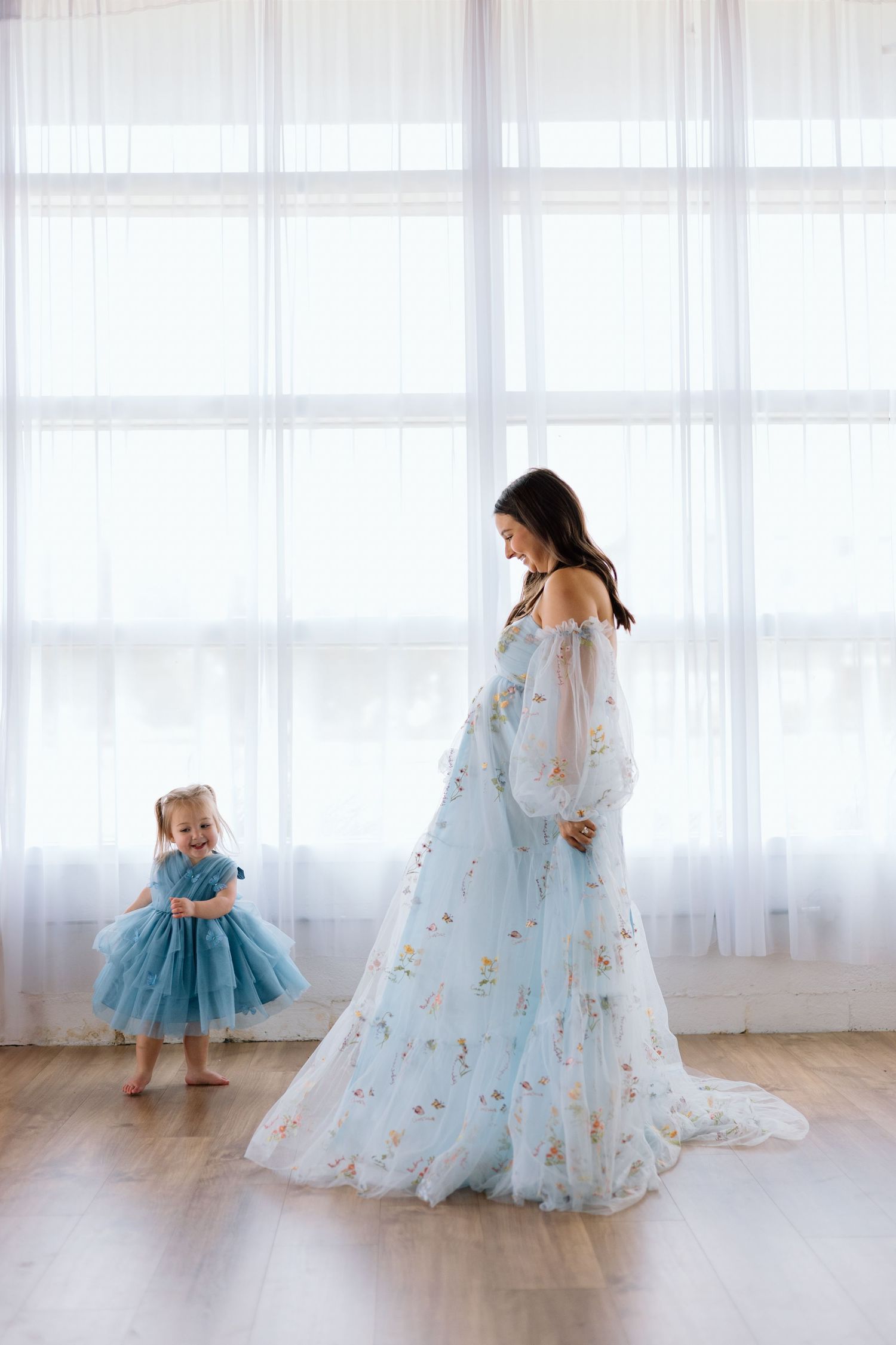 Mother and daughter in matching light blue dresses pose together in a bright, airy room with sheer curtains.