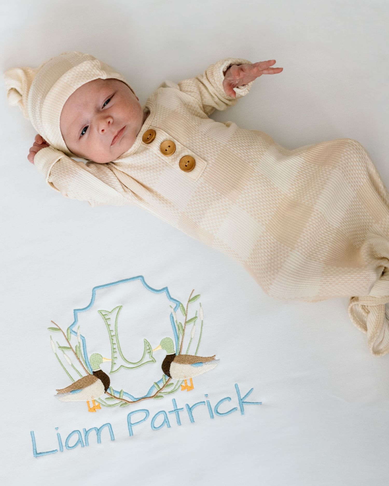 Newborn baby in cream knit gown and hat beside personalized white blanket embroidered with Liam Patrick and duck crest.