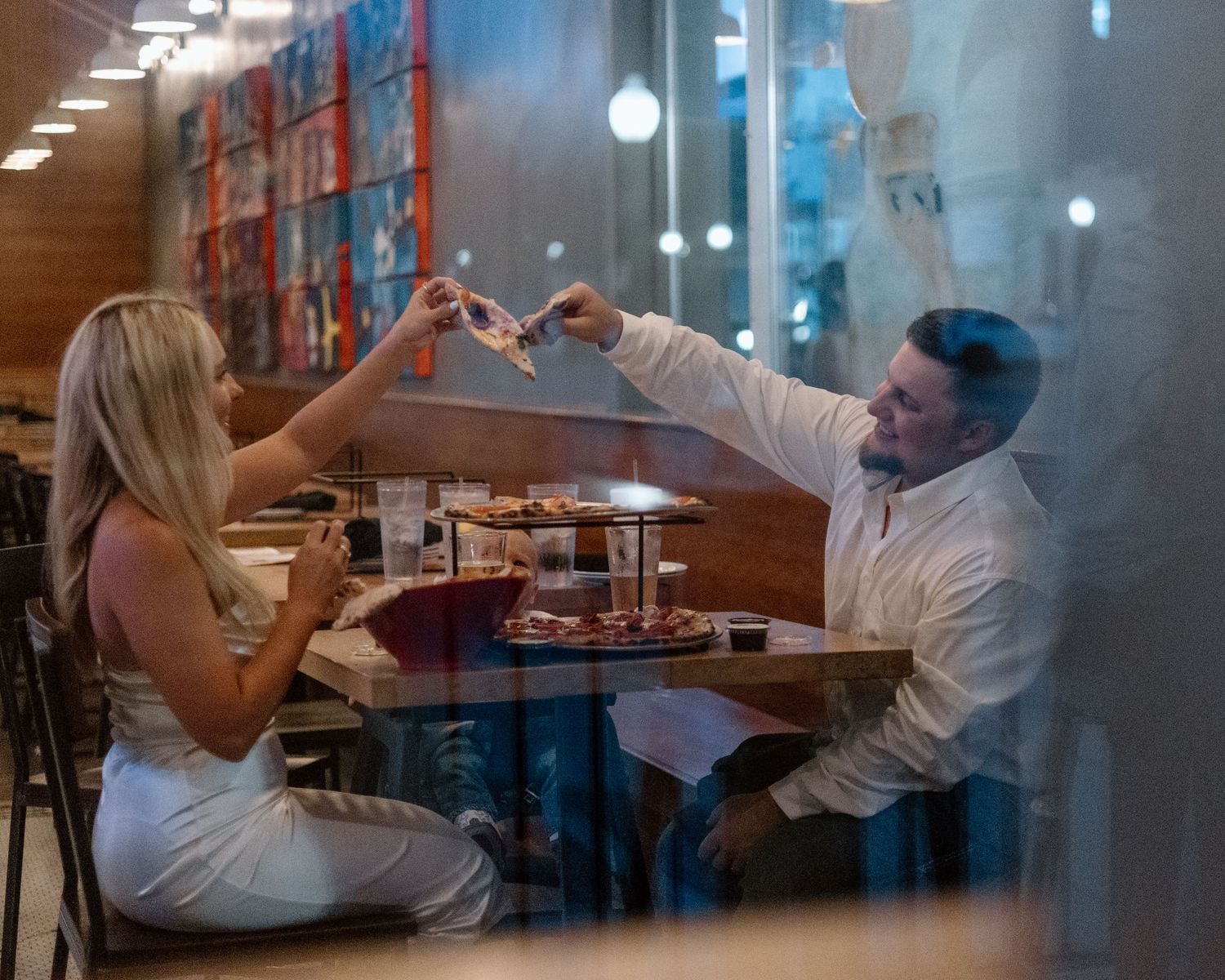 Couple toasting pizza slices at a restaurant table, enjoying a romantic dinner date indoors.