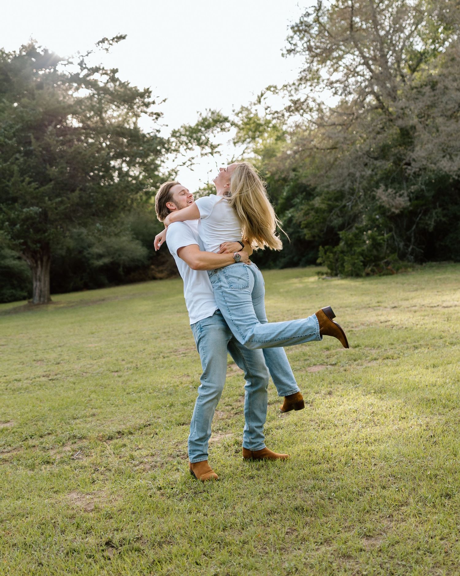 A couple joyfully embraces in a sunny green park, with the man lifting the woman as they share a romantic moment.