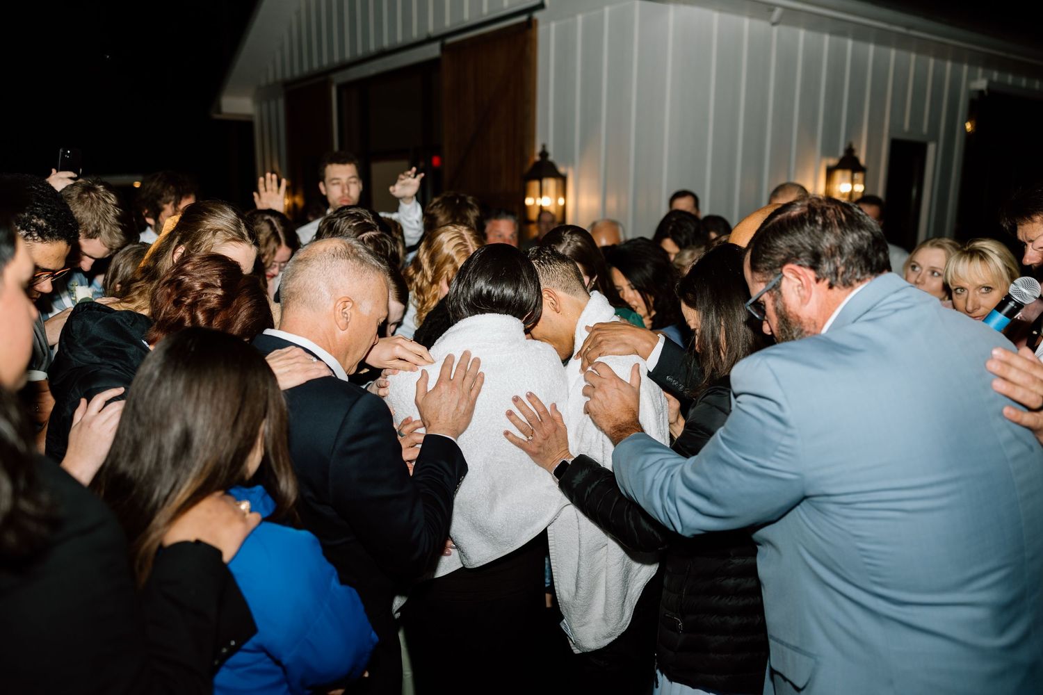 Wedding guests surrounding and praying over newlyweds during outdoor reception celebration.