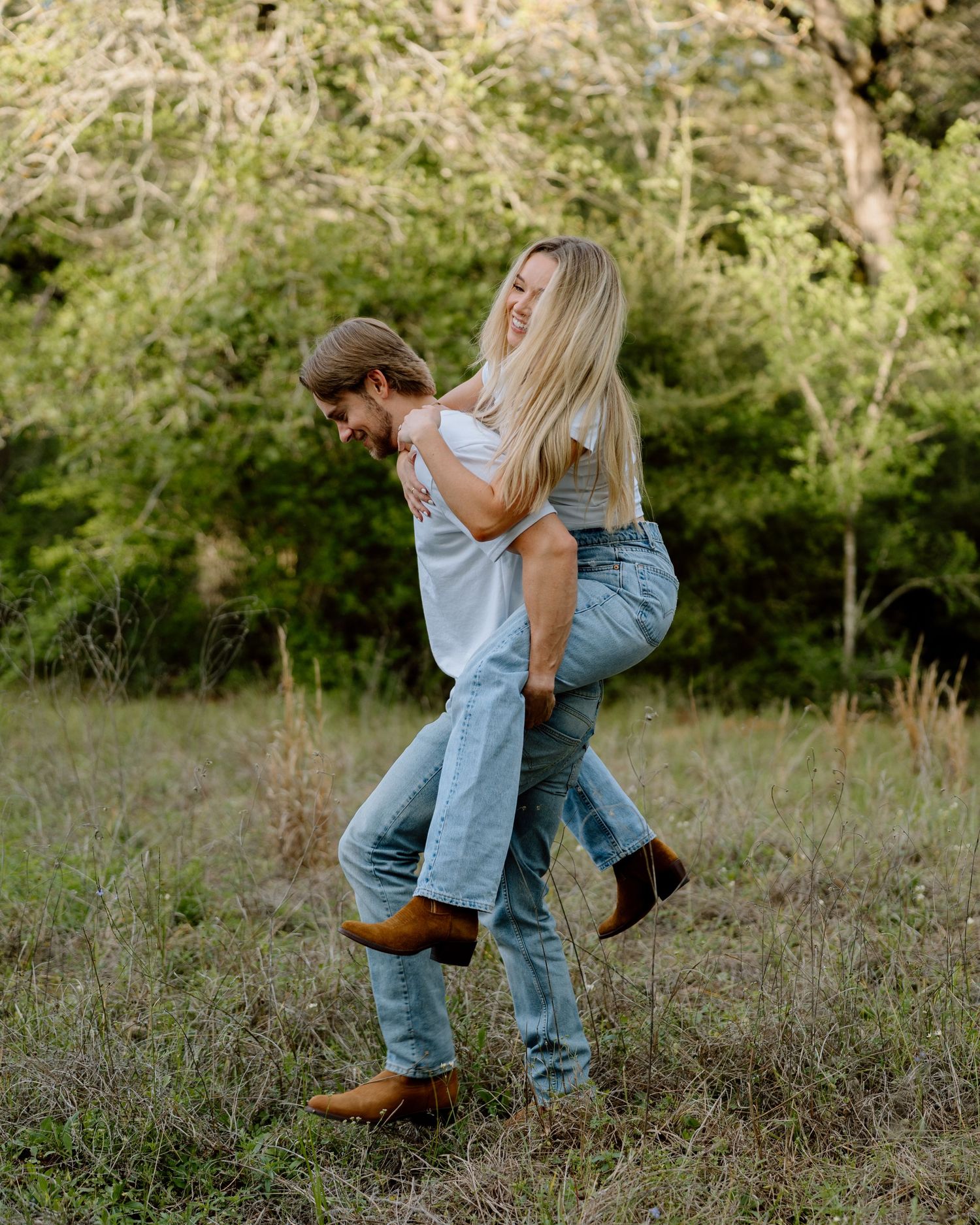 Man giving laughing woman a piggyback ride through a grassy field with trees in background.