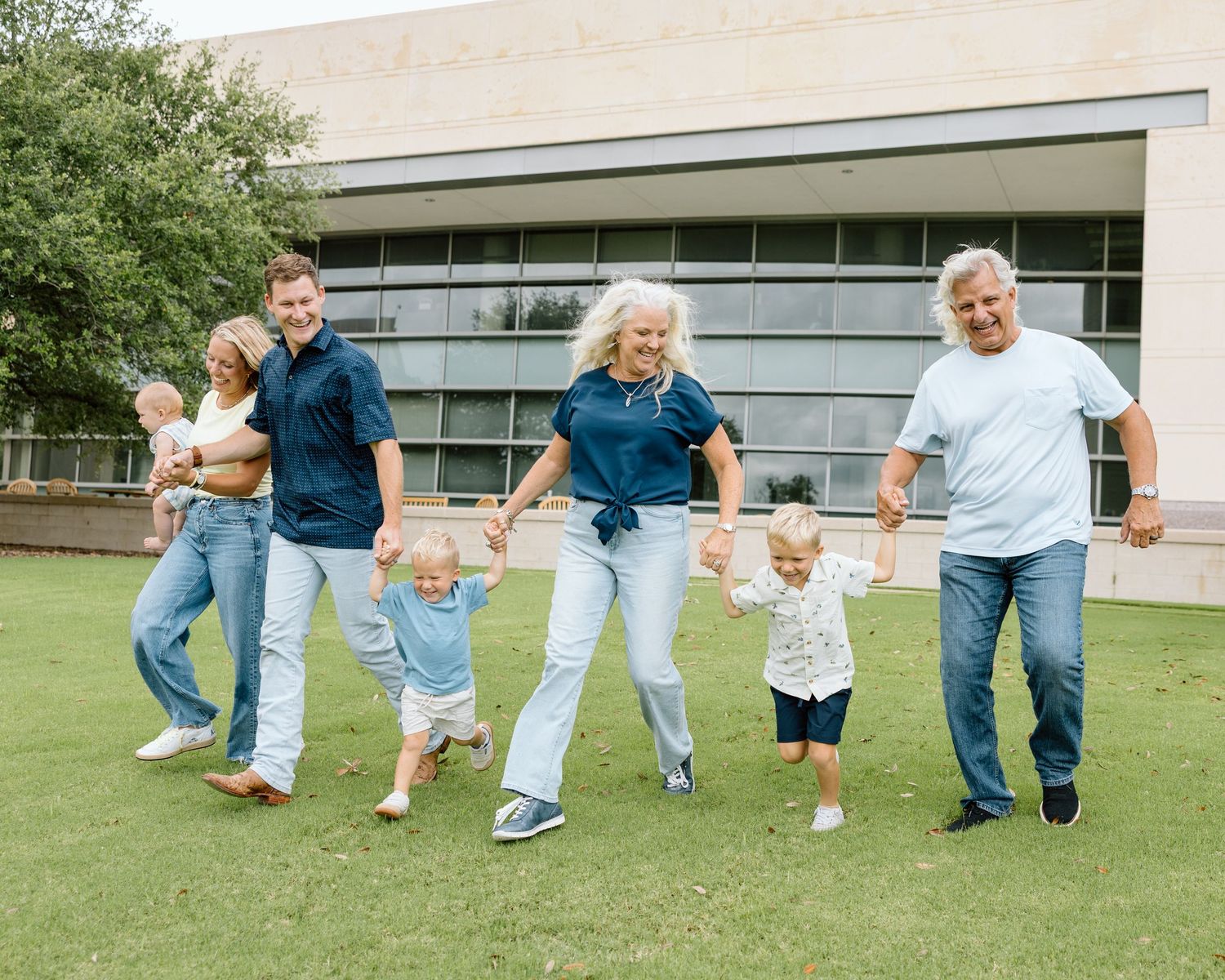 Multi-generational family holding hands and walking together on green grass outside a modern building.
