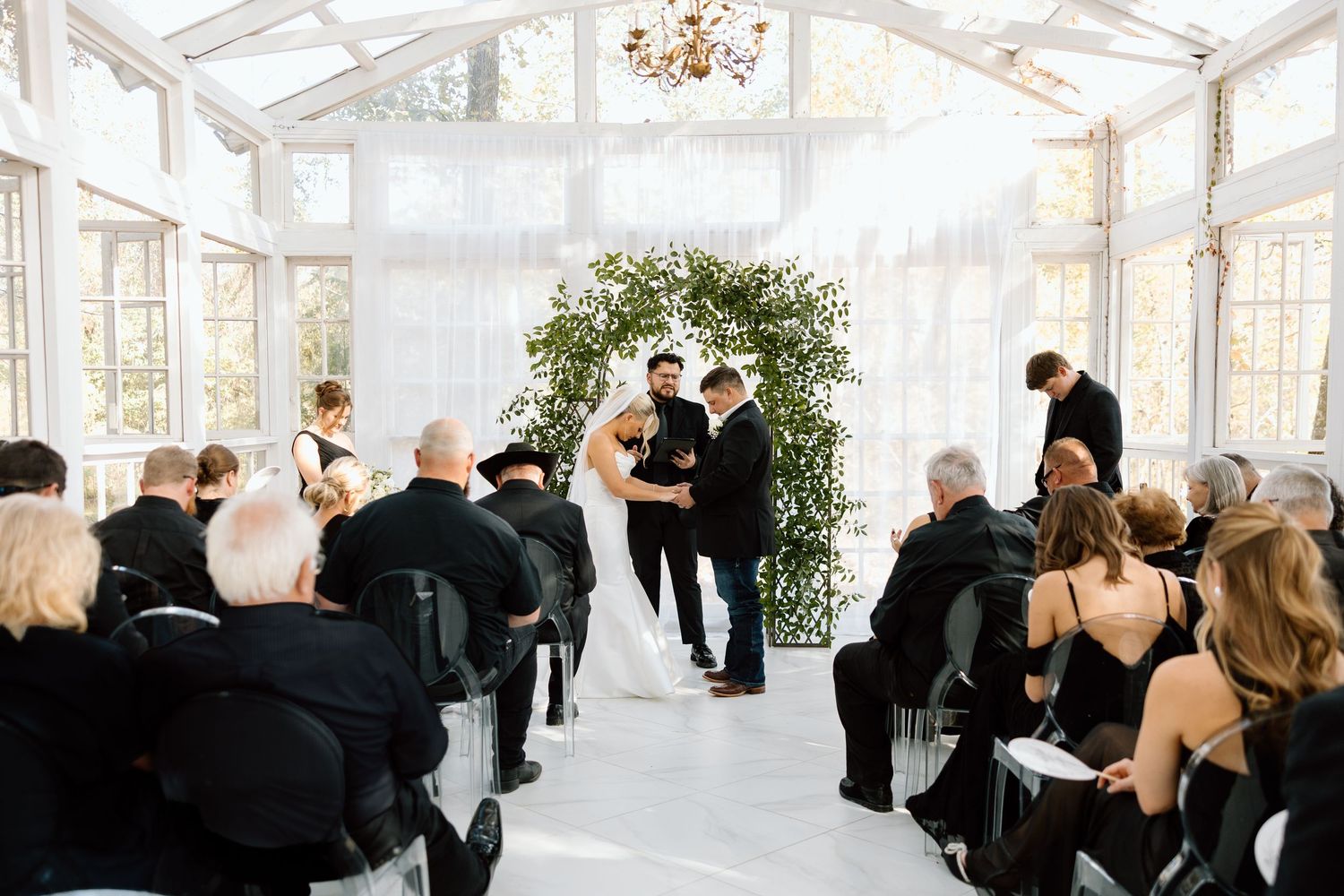Wedding ceremony inside white greenhouse venue with guests seated in black attire watching couple exchange vows.