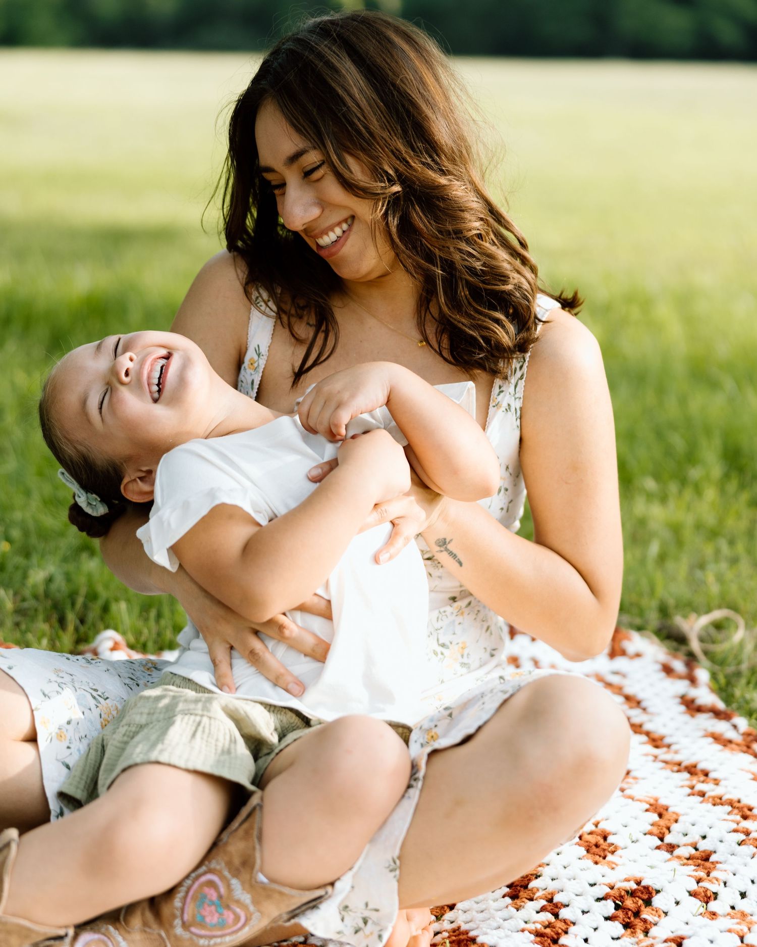 A smiling mother sits on a blanket in a green field, playfully cradling her laughing young daughter.