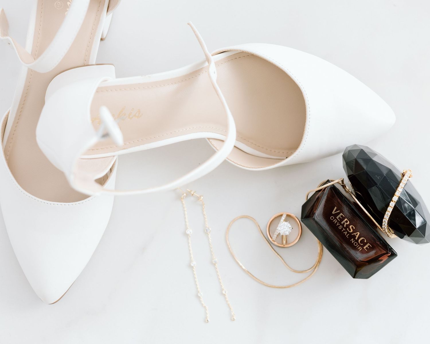 White bridal heels, gold pearl earrings, and Versace perfume flatlay on white surface.