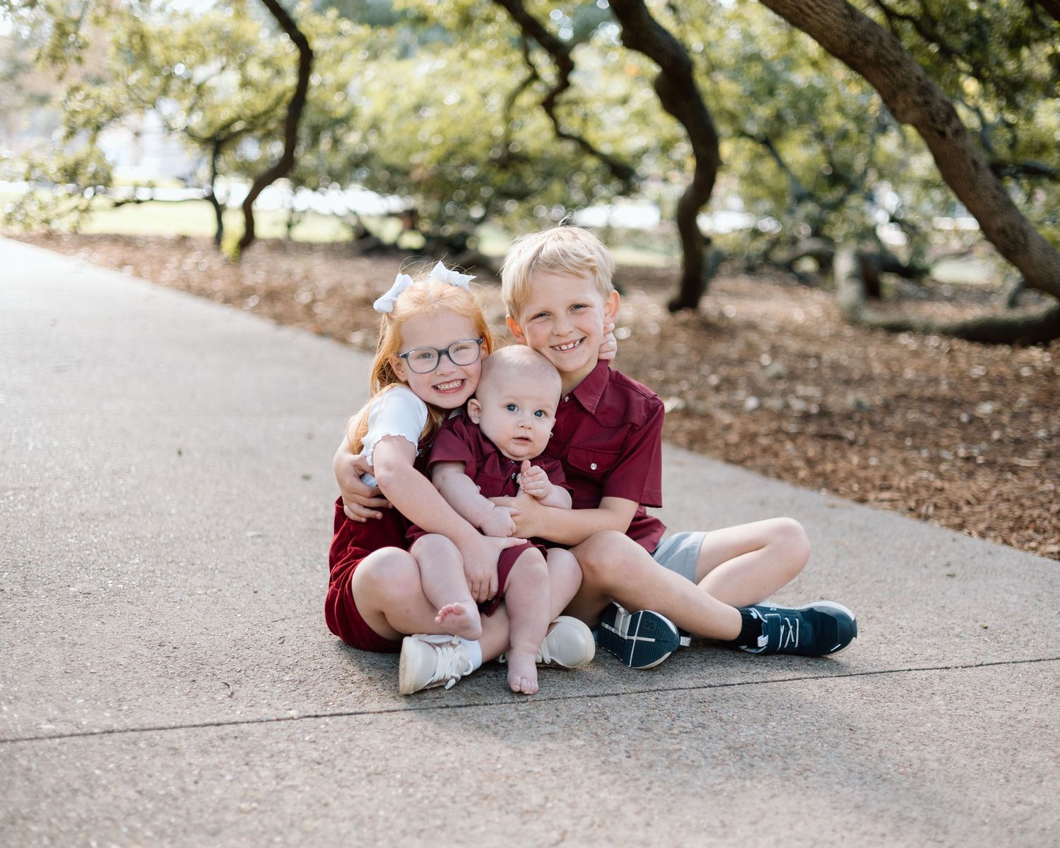 Three young siblings sit together on a path outdoors, smiling and hugging, dressed in matching maroon outfits.