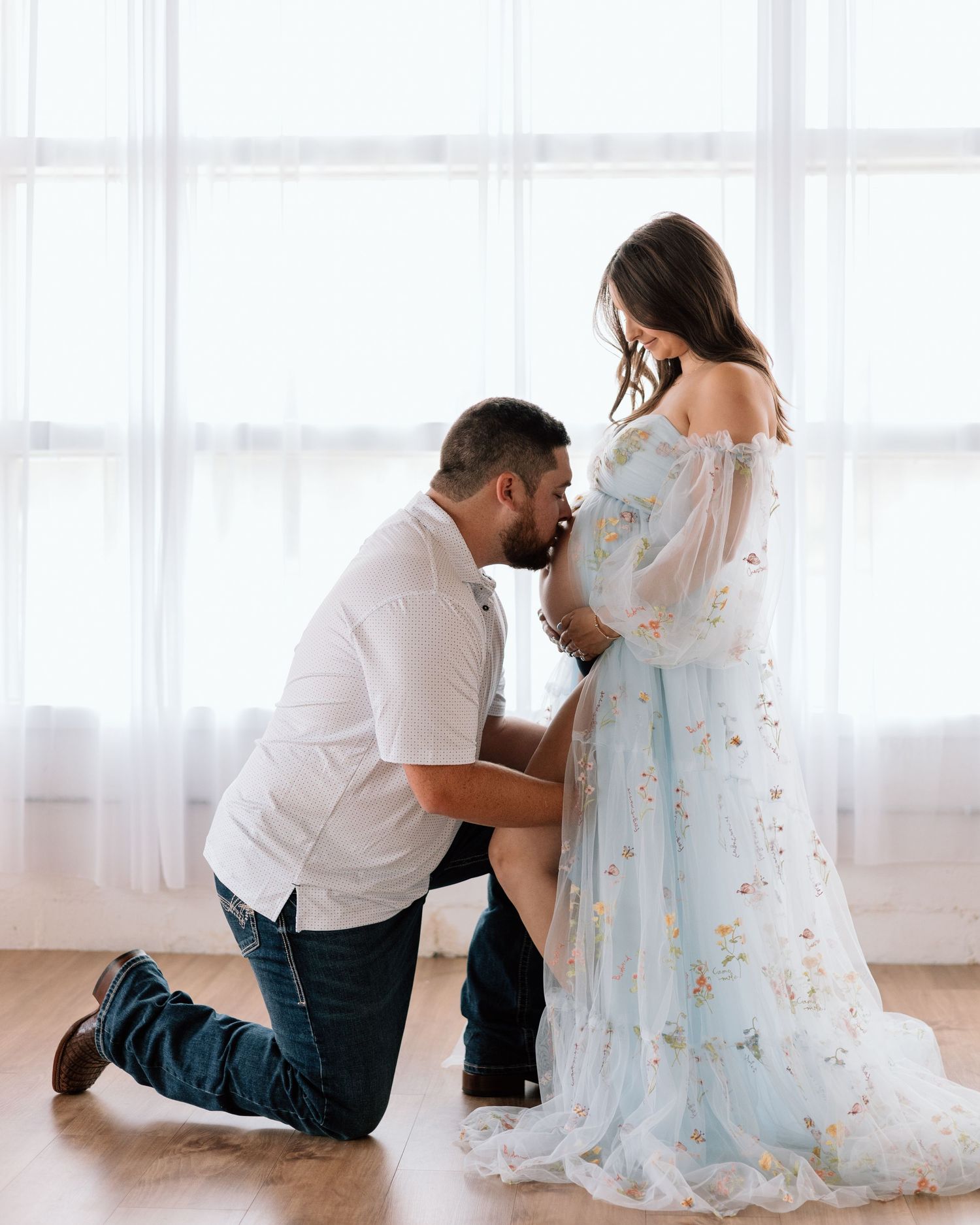A man kneels kissing a pregnant woman's baby bump during a maternity photo session in a light-filled studio.