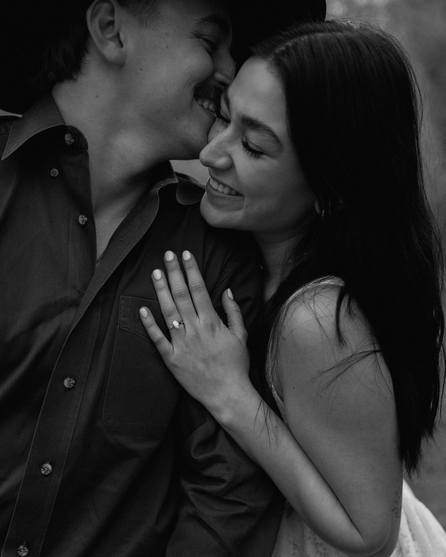 Black and white photo of a smiling couple sharing a romantic moment, woman displaying an engagement ring on her hand.