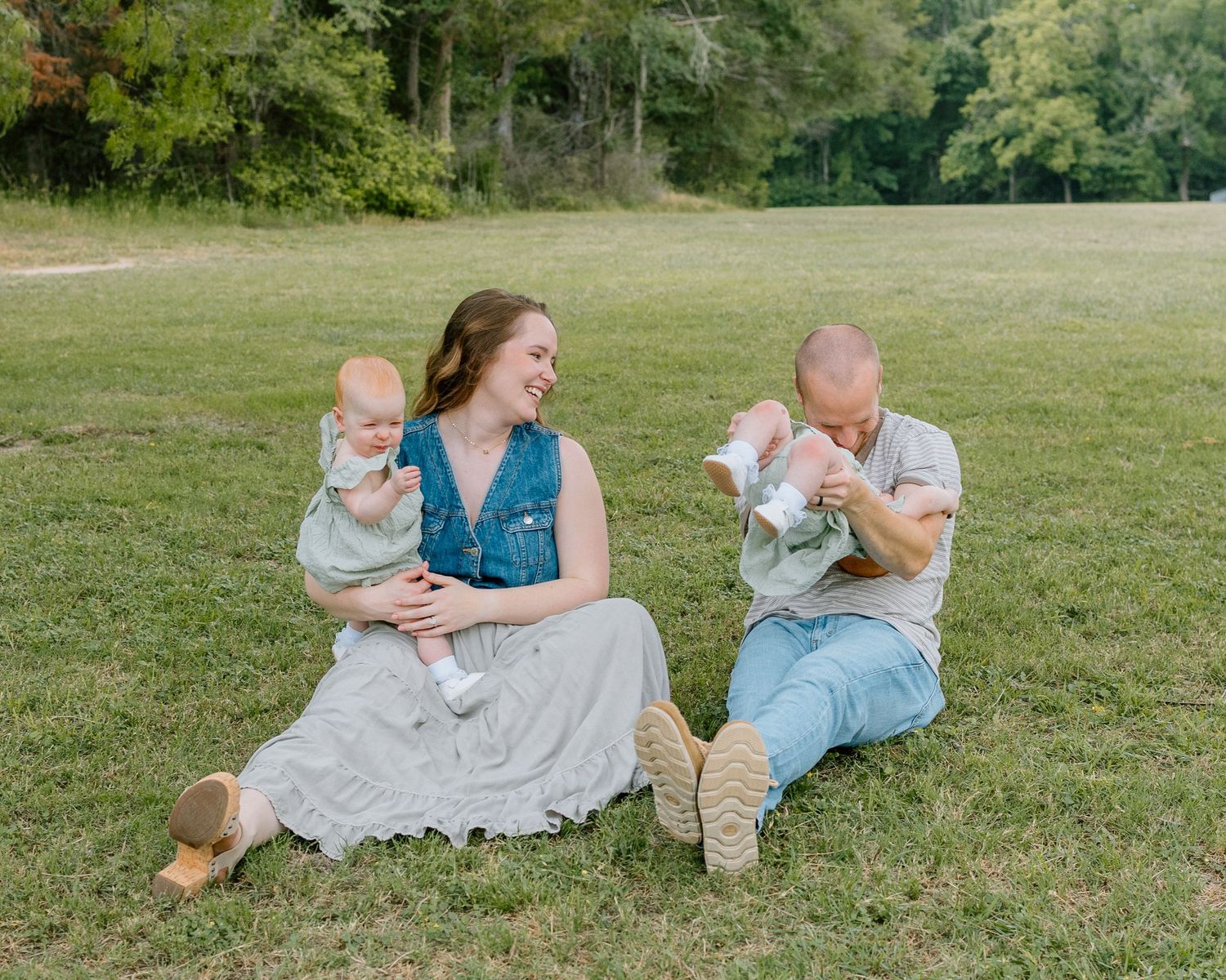 A couple sits in a grassy field on a sunny day, each holding an infant while wearing casual summer attire.