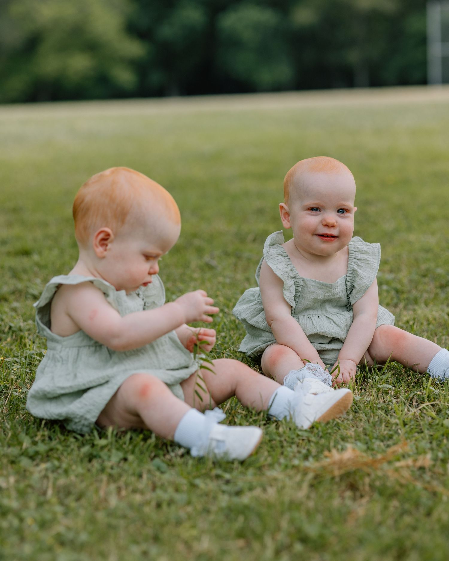 Two babies in matching mint green rompers sit together on grass on a sunny day while wearing white socks.