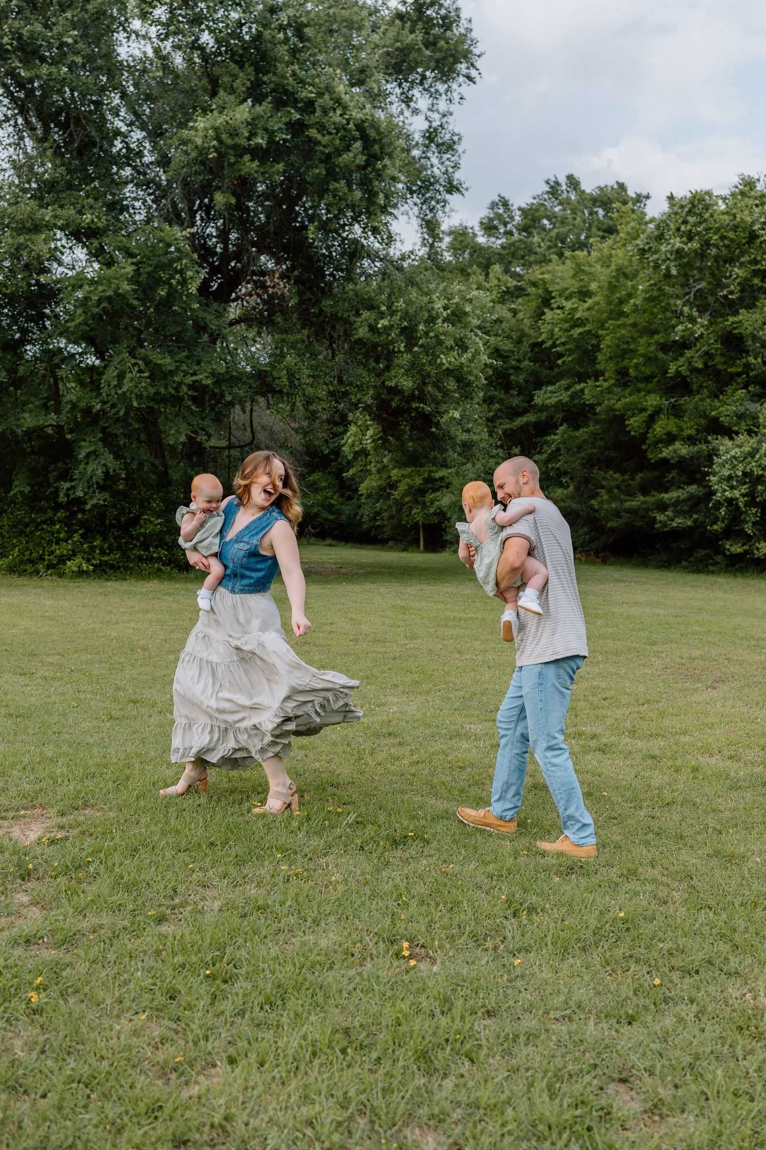 Couple dancing and twirling with babies on a grassy lawn surrounded by leafy green trees on a summer day.
