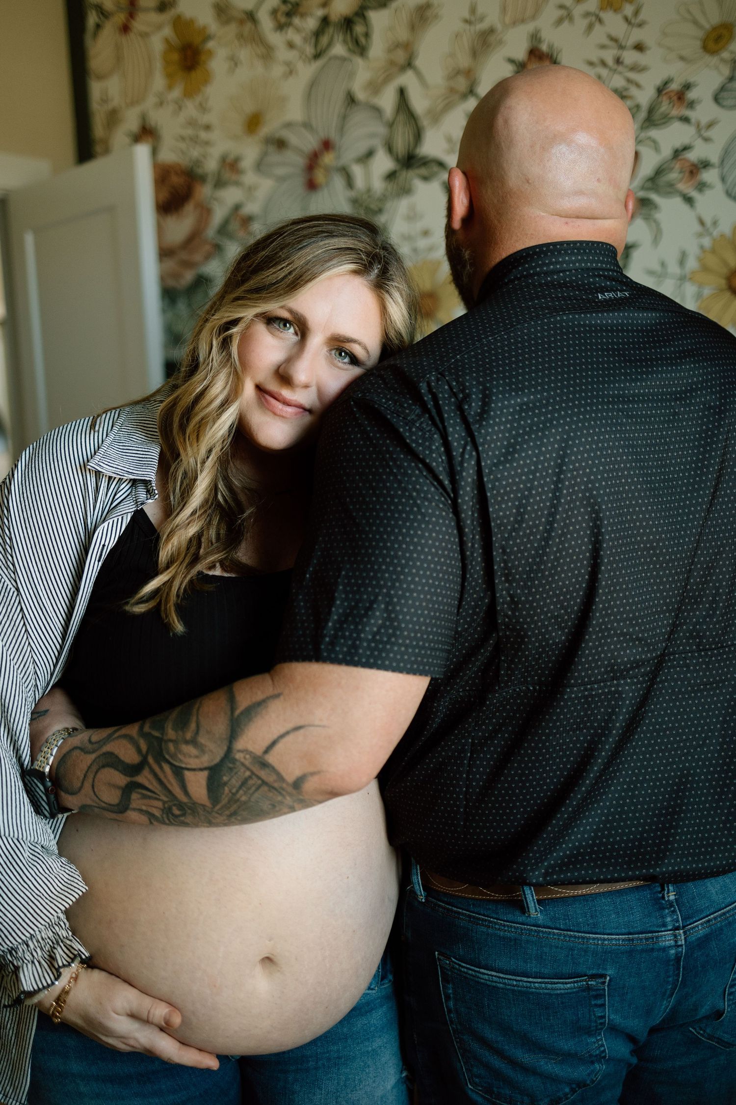 Maternity photo with couple in casual attire posing against floral wallpaper backdrop.