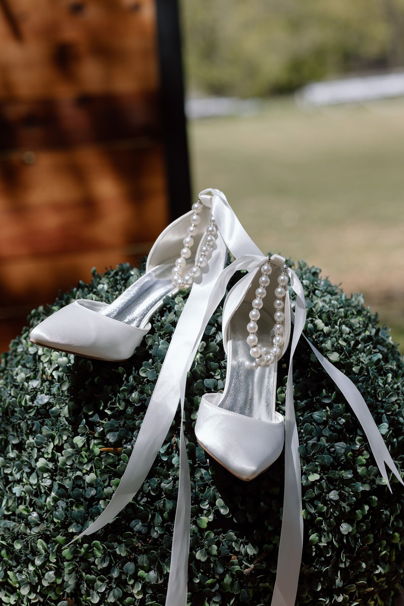 Elegant white bridal shoes with pearl straps displayed on a decorative green topiary bush.