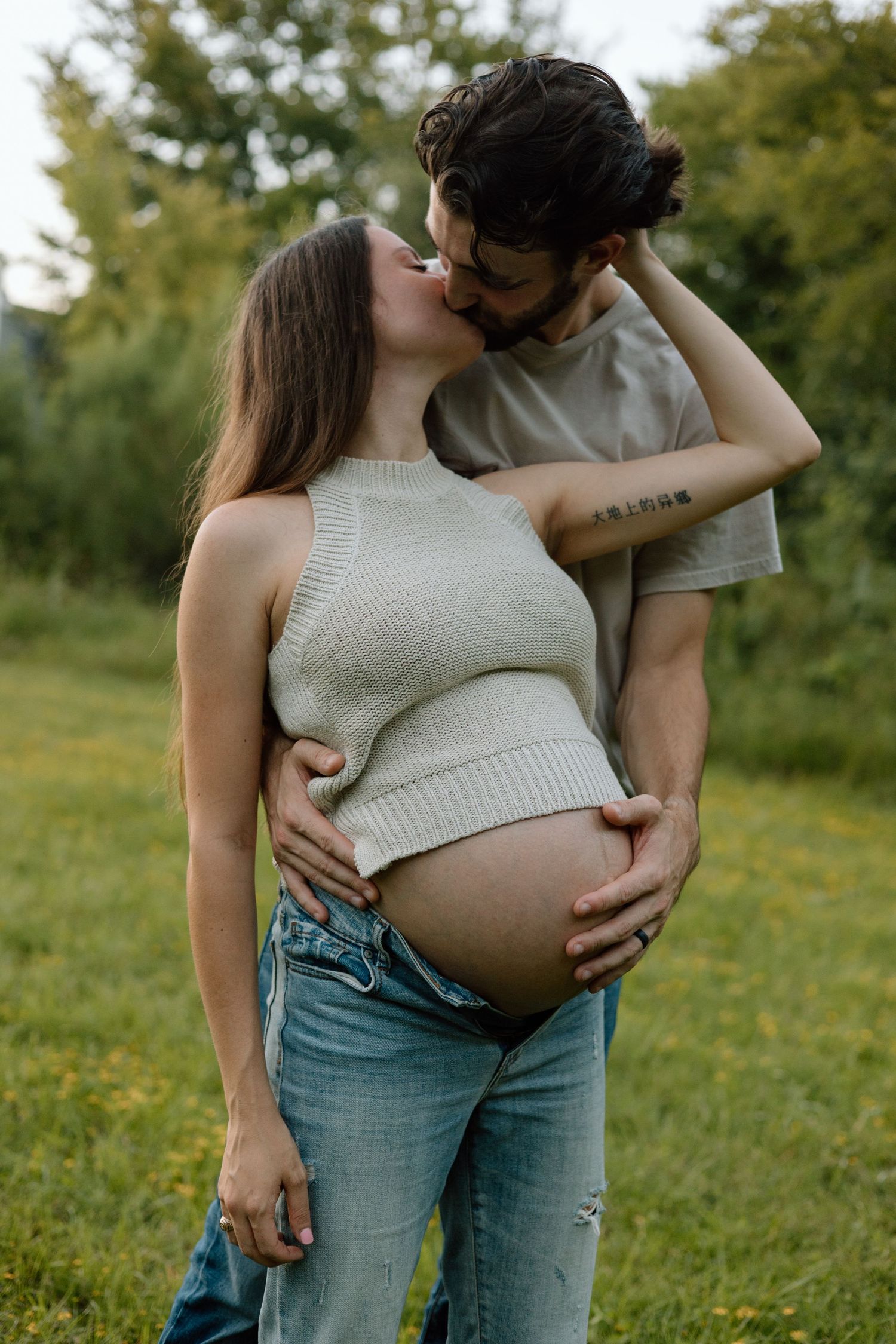 Intimate maternity photo of a couple in casual wear standing in a sunlit garden.