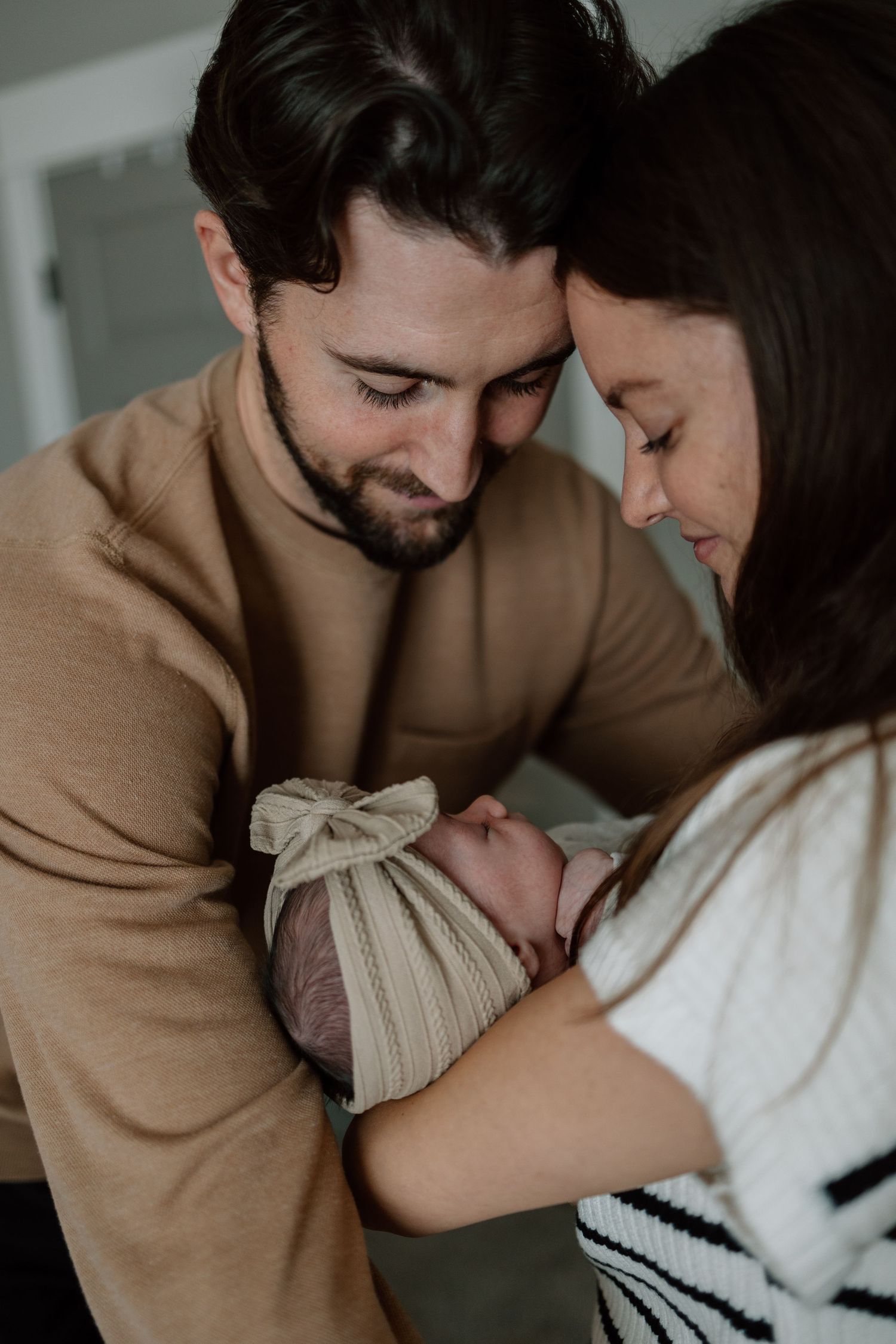 Tender family moment with newborn baby wearing beige bow headband held between parents.