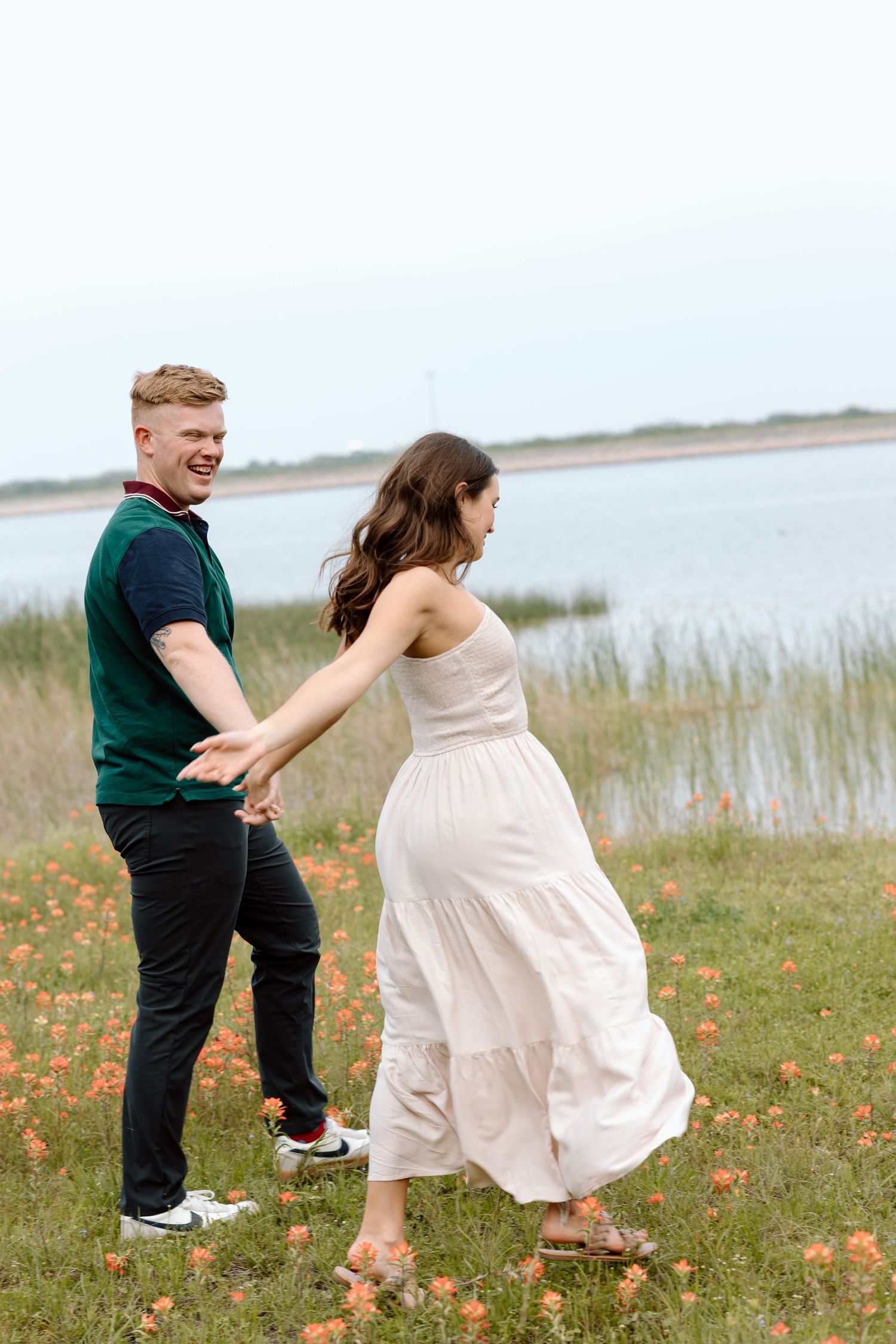 A couple holds hands while dancing through wildflowers near a serene lake.