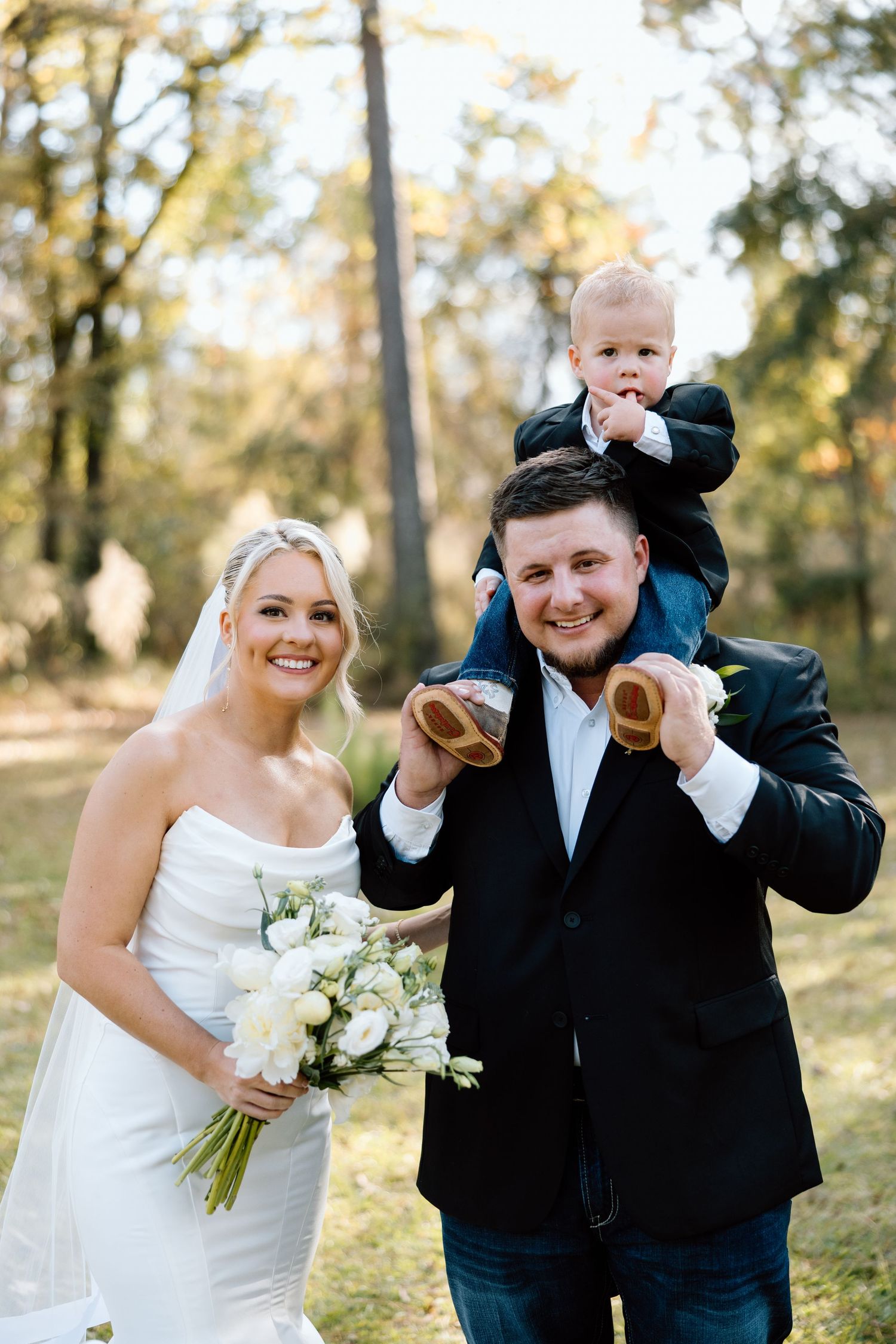 Smiling bride and groom posing outdoors with a baby on grooms shoulders holding tiny shoes.