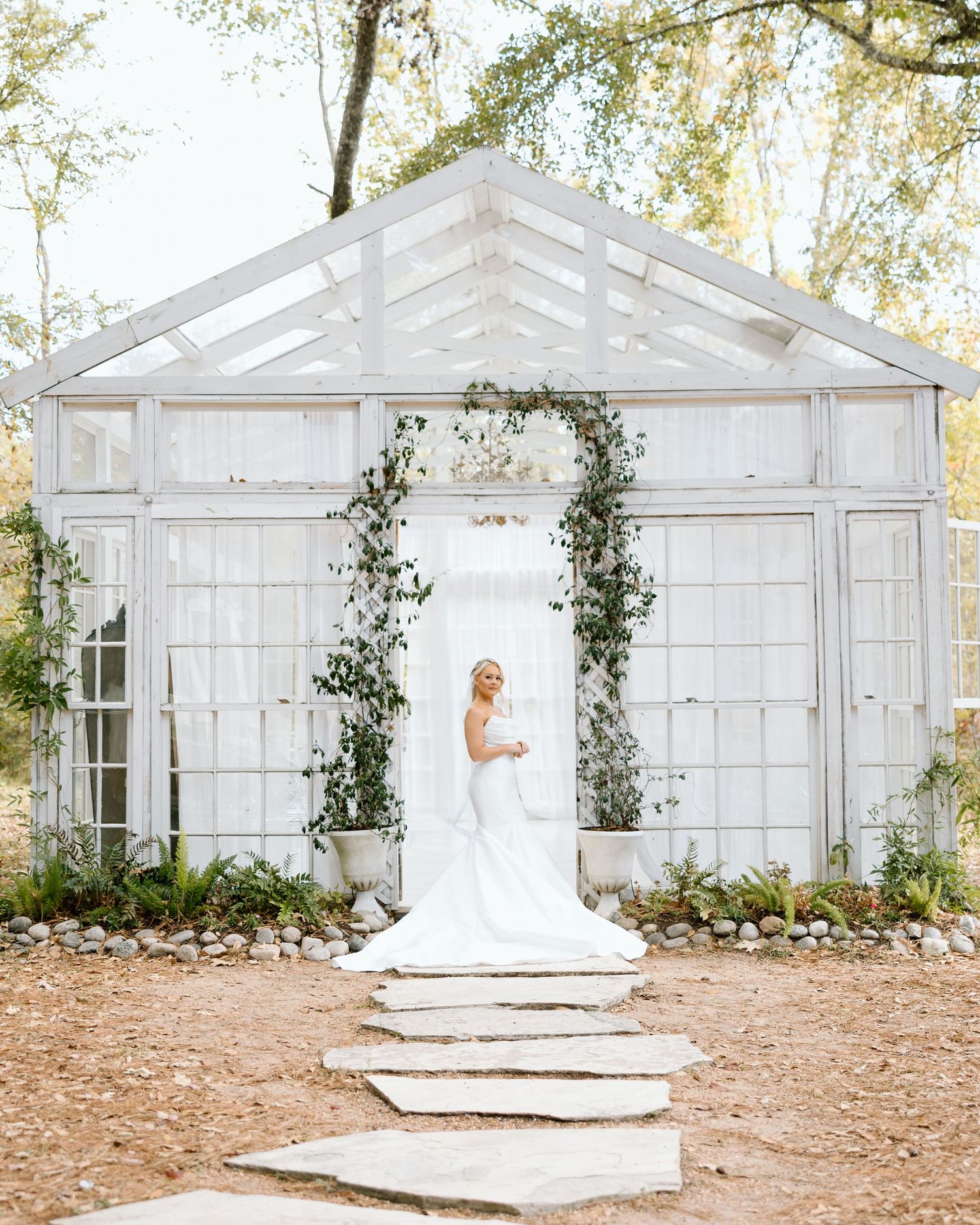 Bride in white mermaid gown poses at entrance of elegant white greenhouse wedding venue with floral arch.