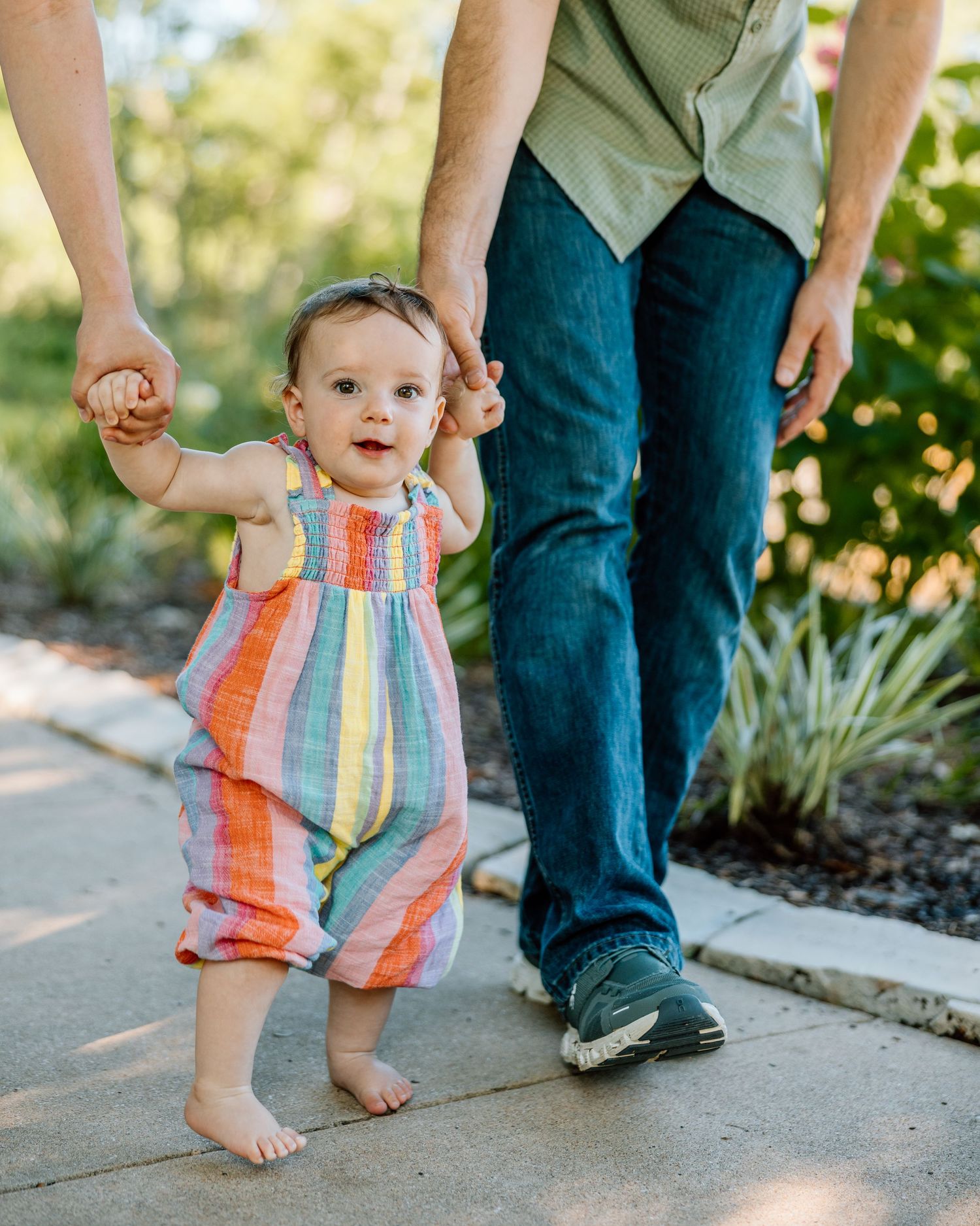 Smiling baby in colorful striped romper takes first steps outdoors, held by adult hands on a sunny garden path.