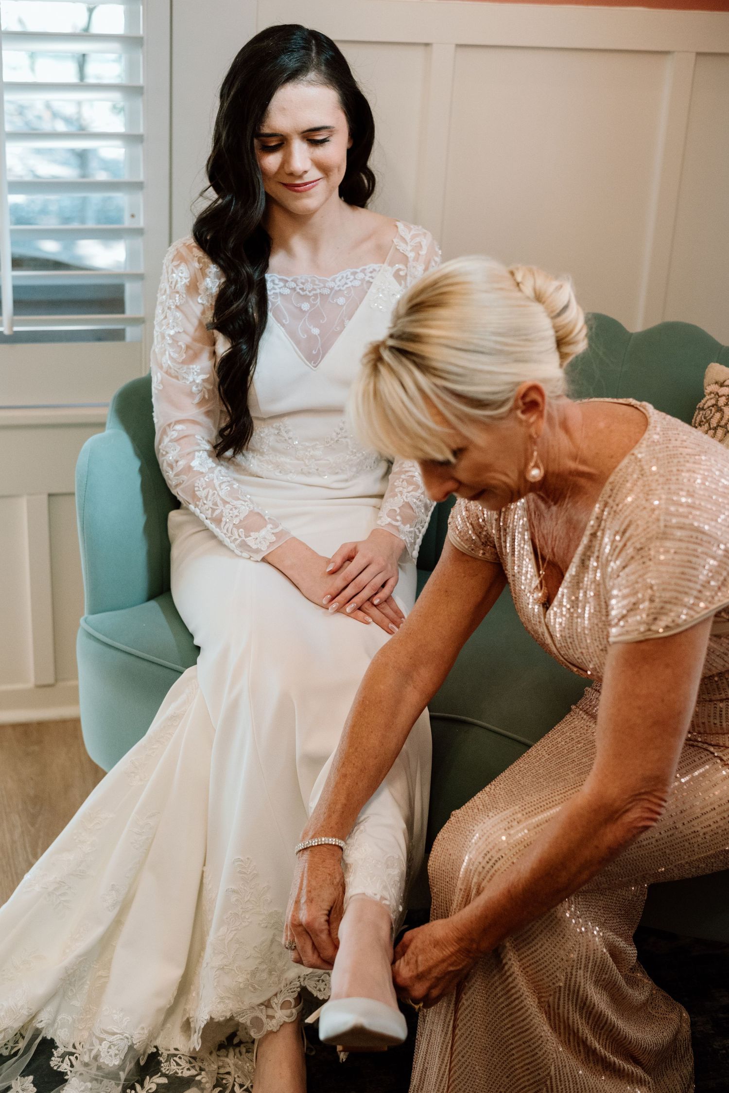 Bride in lace wedding gown smiles as older woman helps her put on white heels while seated on teal chair.