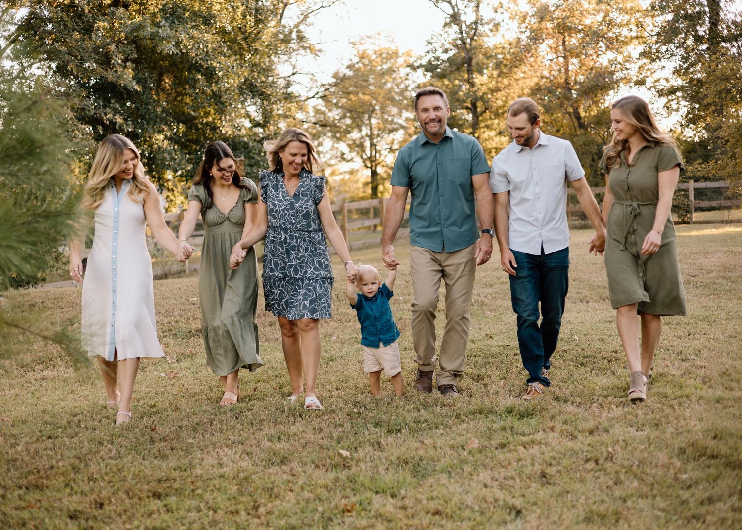 Happy family of six walking together outdoors in a sunny park, smiling and holding hands in casual attire.