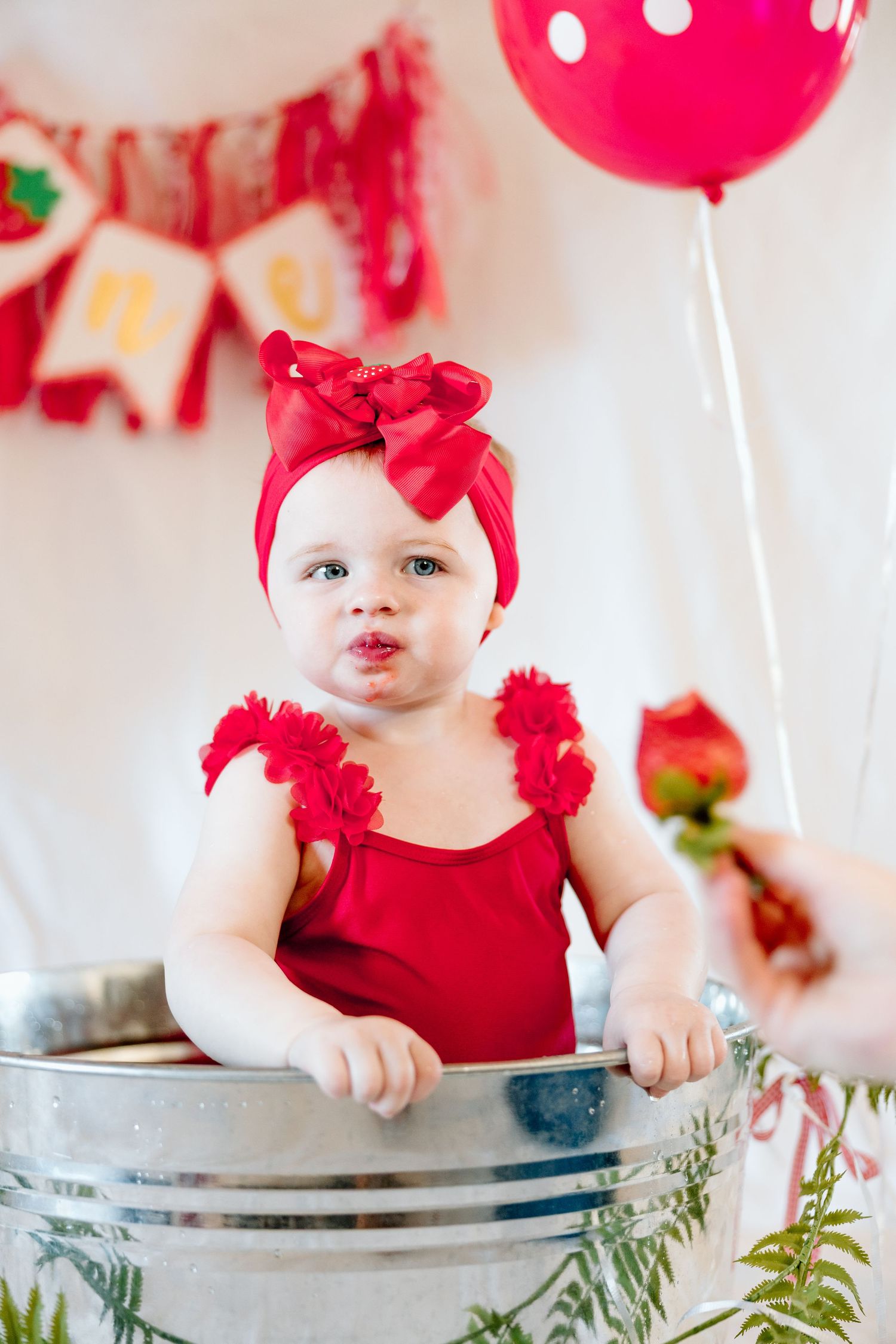 Baby girl in red romper and bow headband sits in metal tub at strawberry first birthday party.